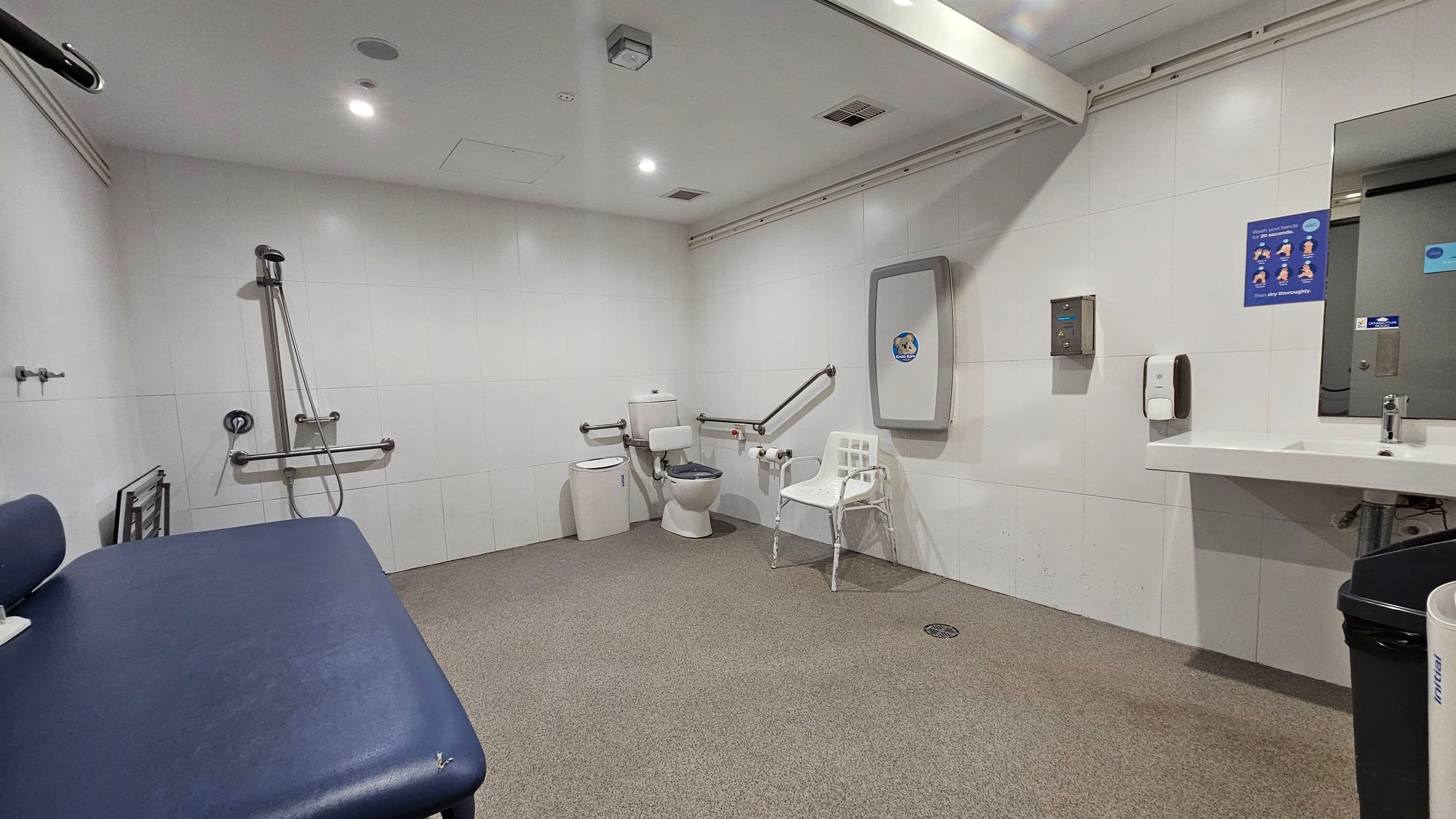A spacious, accessible bathroom with white walls and a beige floor. On the left, there's a blue padded changing table and a shower area with grab bars. In the centre, a toilet with support rails and a white plastic chair nearby. On the right, a sink with a mirror above, a soap dispenser, and a hand dryer. A baby changing station is mounted on the wall. The ceiling is white with recessed lighting.