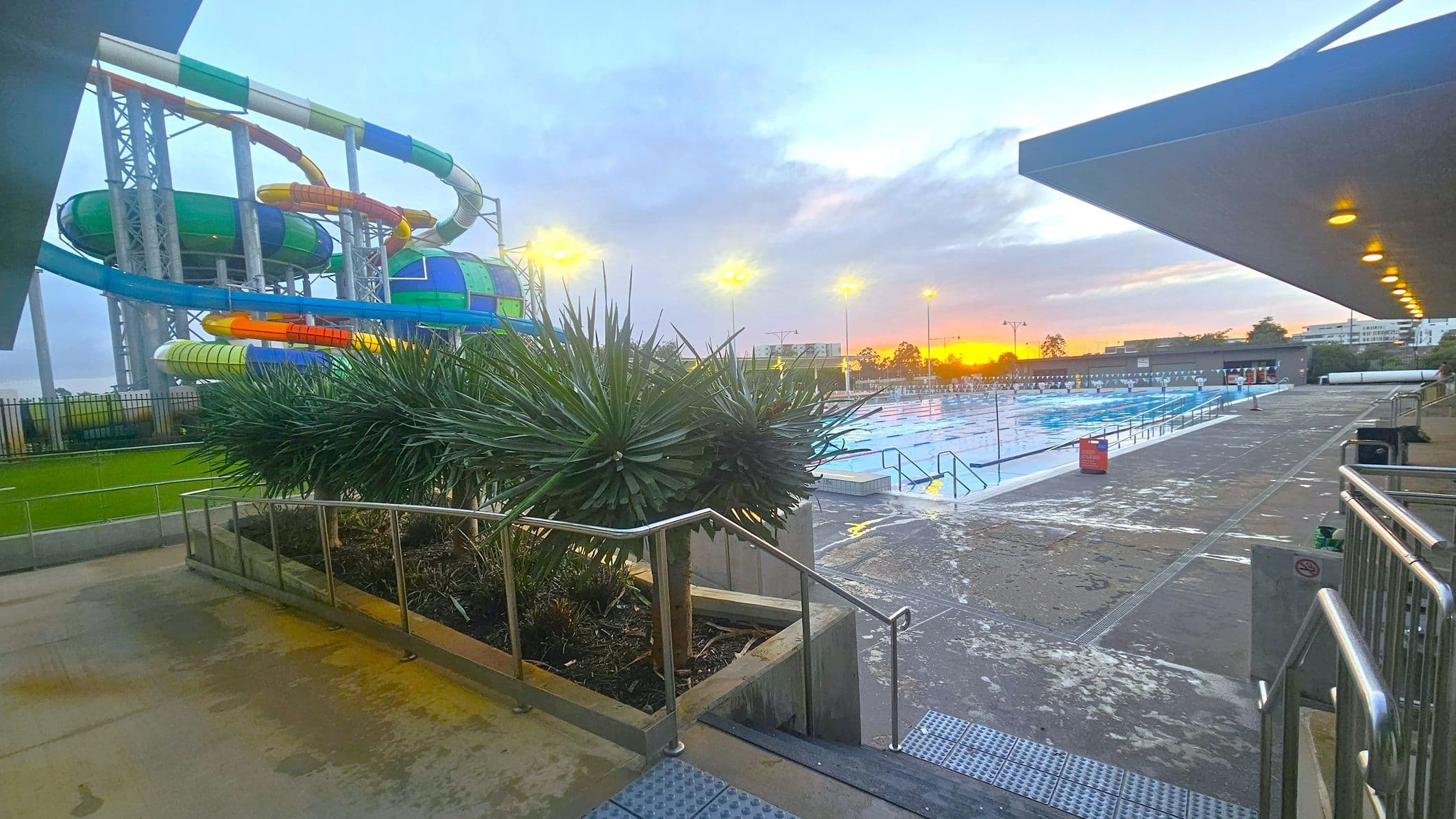On the left, there are colourful water slides with a mix of green, blue, orange, and yellow sections supported by metal structures. In the middle, spiky green plants are bordered by metal railings. On the right, a large outdoor swimming pool is visible, with lane dividers and a wet concrete deck. Overhead, a roof with recessed lighting extends partially over the pool area. In the background, the sun is setting, casting a warm glow over the scene.