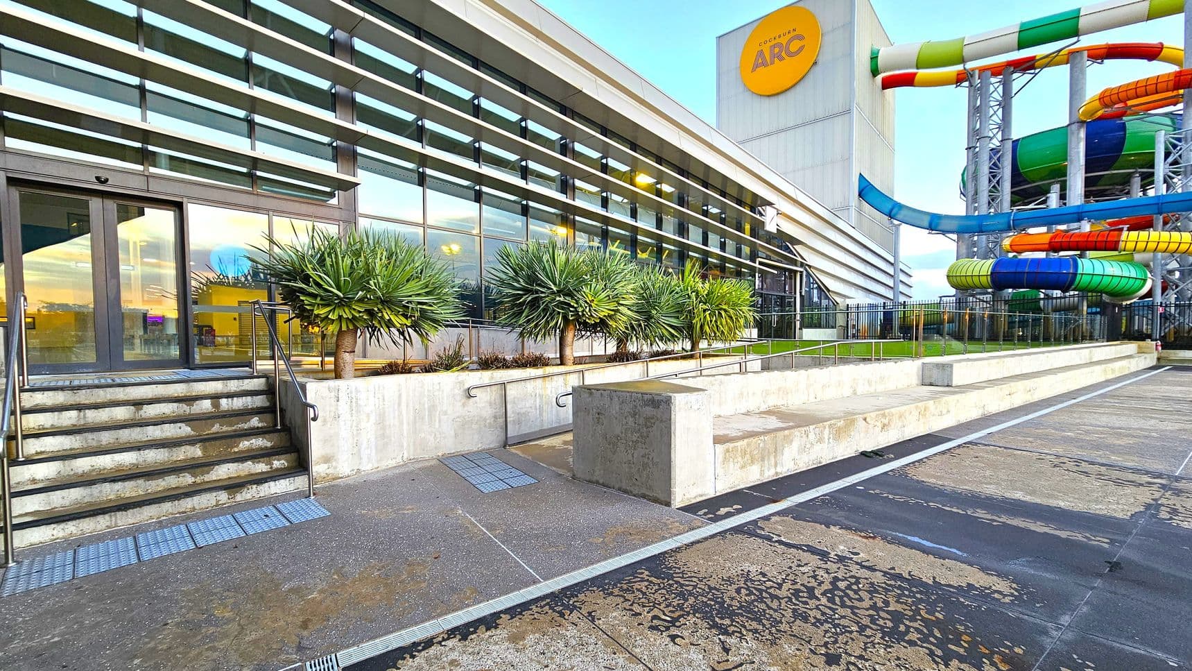 A modern building with large glass windows on the left, featuring a set of stairs leading to an entrance. Several spiky-leaved plants are lined up along the building. On the right, colourful water slides in green, blue, orange, and yellow are visible, supported by metal structures. The ground is a mix of concrete and textured surfaces, and a low concrete wall runs parallel to the building. A sign with "ARC" is mounted on the building.