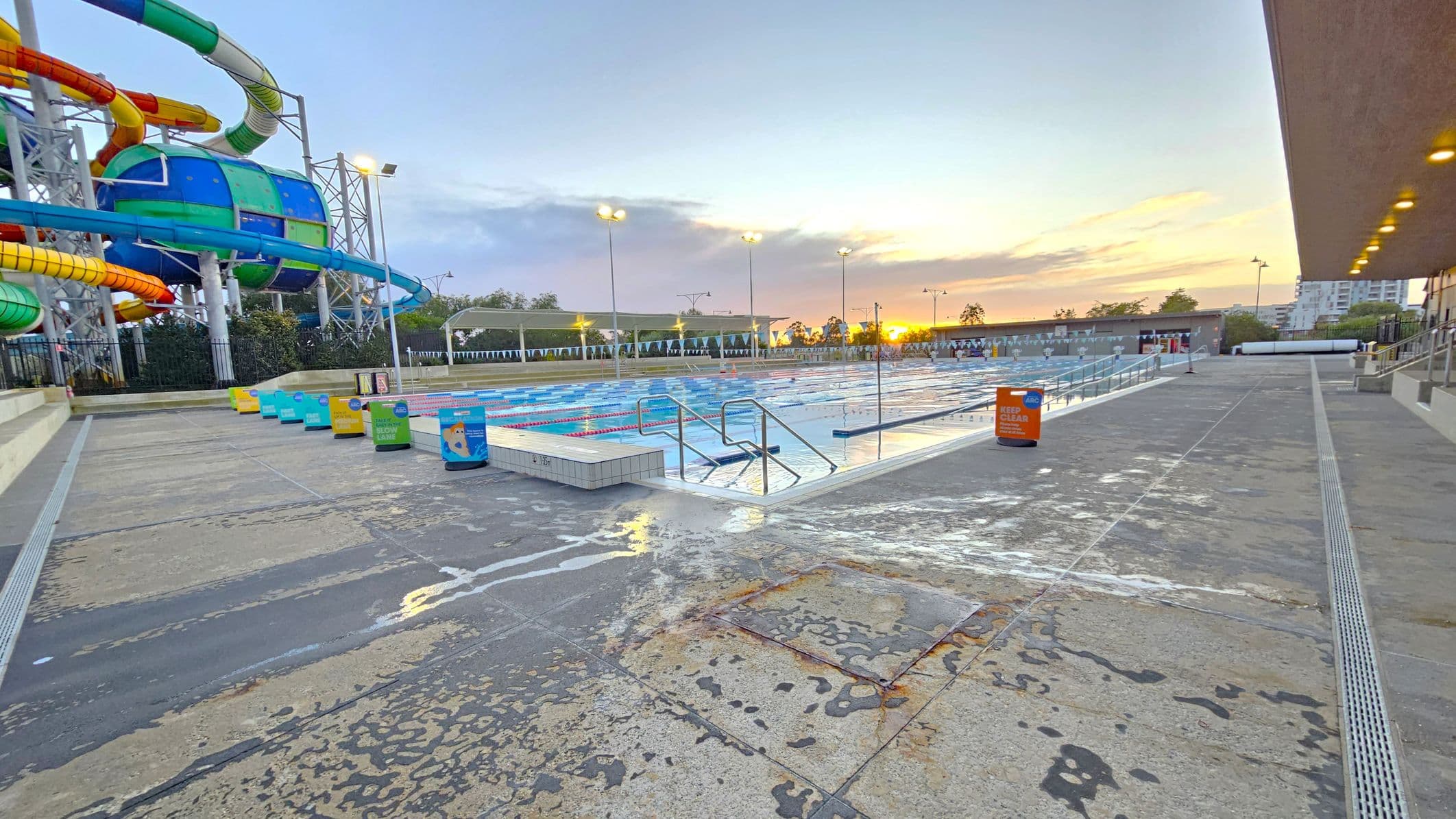 On the left, there is a colourful water slide structure with multiple twisting tubes in green, blue, yellow, and orange. In the centre, a large outdoor swimming pool with lane dividers is visible, surrounded by a wet concrete deck. Several bins with colourful labels are lined up along the poolside. On the right, a building with a flat roof and lights underneath extends along the pool area. The background shows trees and a sunset sky.