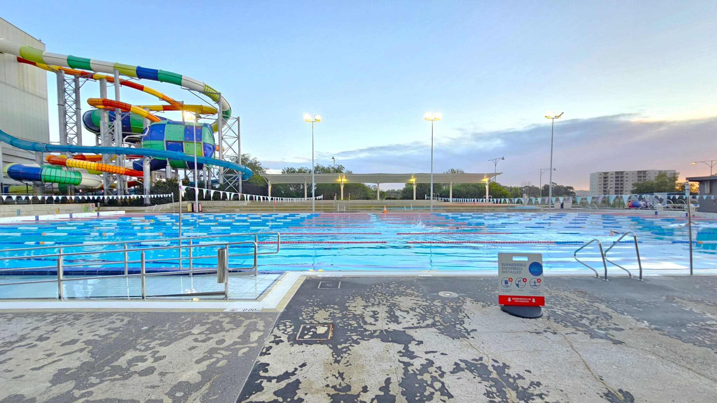 An outdoor swimming pool with multiple lanes is in the centre, surrounded by a concrete deck. On the left, there is a colourful water slide structure with twists and turns. In the background, tall light poles illuminate the area, and there are trees and a few buildings visible. A sign with safety instructions is placed near the pool's edge on the right. The sky has a gradient of blue and pink hues.
