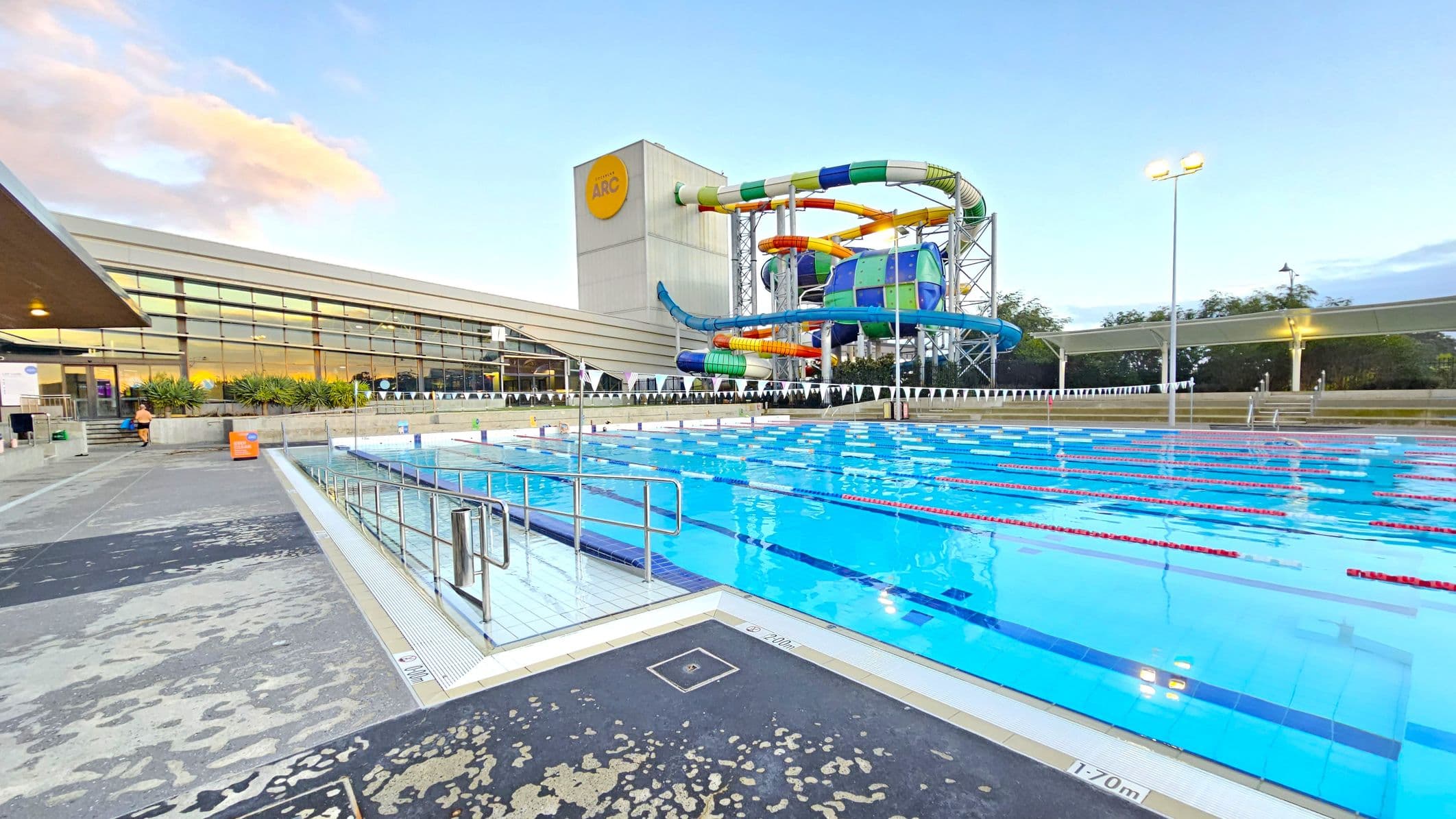 On the left, a modern building with large glass windows and a person walking near the entrance. In the centre, a large outdoor swimming pool with lane dividers and a depth marking of 1.70 metres. On the right, a colourful water slide structure with multiple twisting tubes. The foreground features a wet concrete poolside area with metal railings. In the background, trees and a lit pathway are visible.