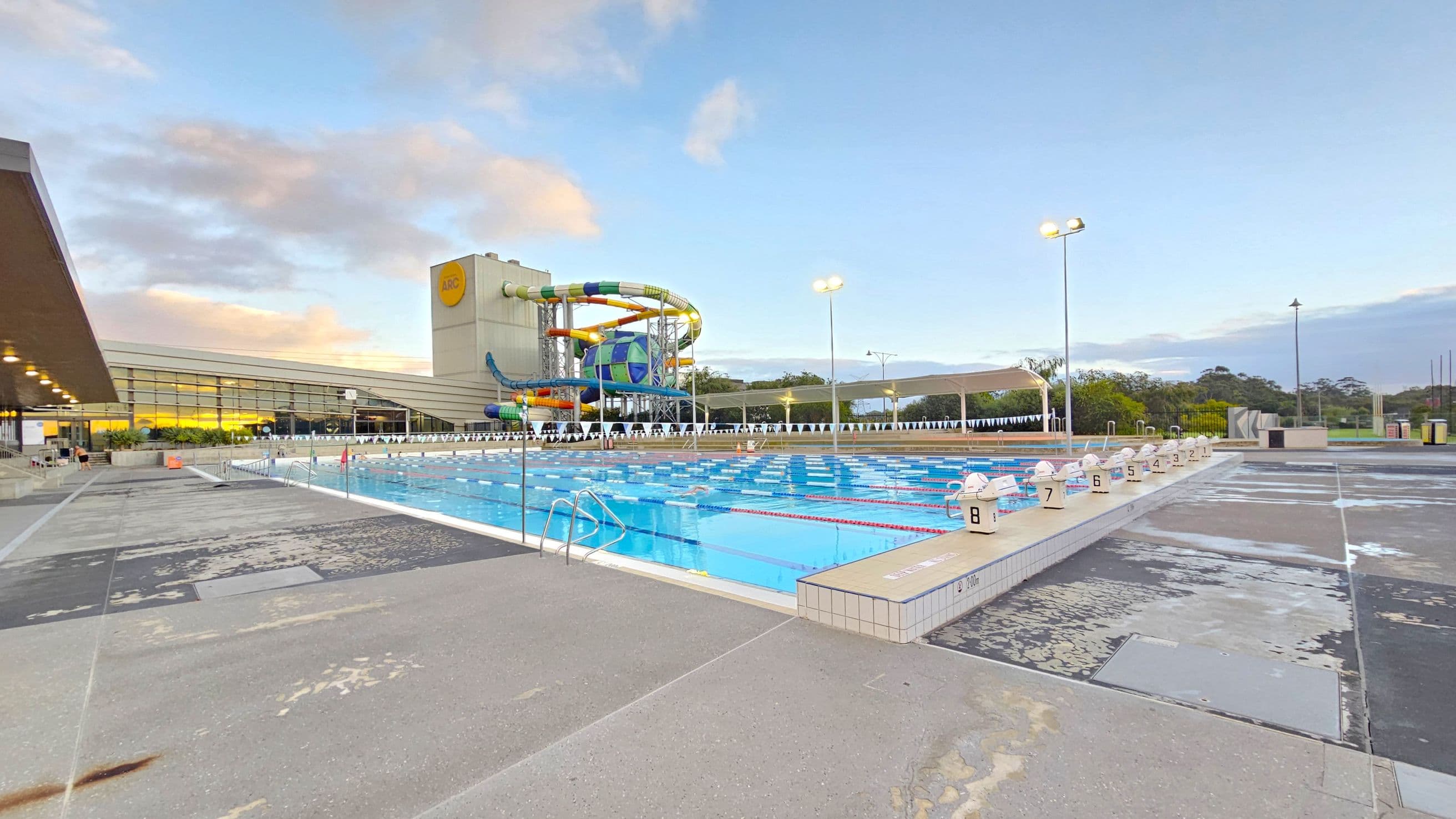 An outdoor swimming pool with multiple lanes is in the centre, surrounded by a concrete deck. On the left, a building with large glass windows and a roof overhang is visible. In the background, a colourful water slide structure is prominent, with a tall tower and twisting tubes. The pool has numbered starting blocks along the edge. Tall lights are positioned around the pool area, and there are trees and greenery in the distance.