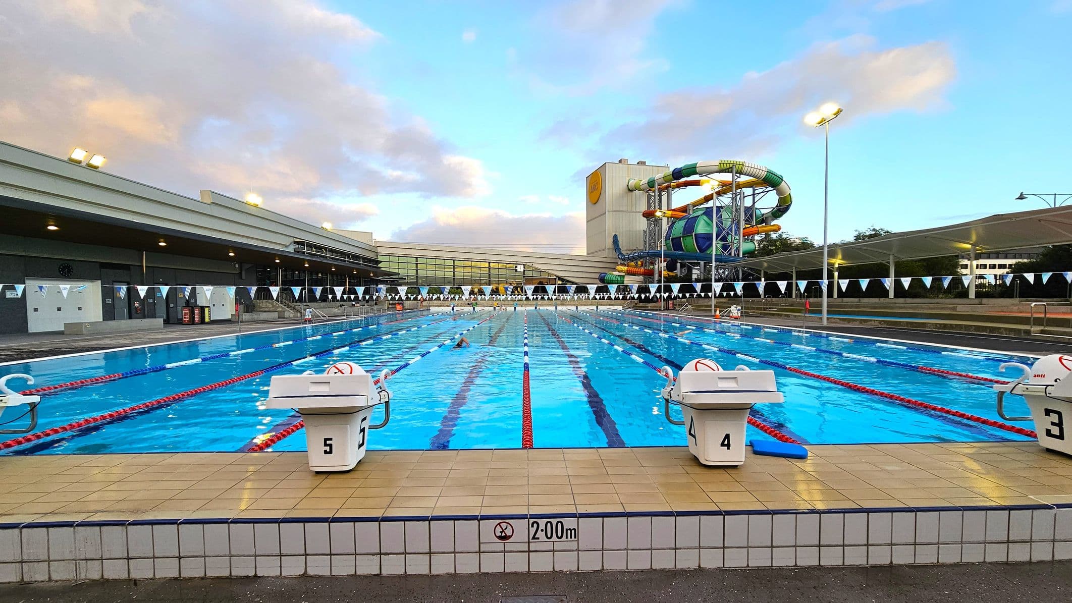 An outdoor swimming pool with clear blue water, divided into lanes by red and blue ropes. Starting blocks numbered 4 and 5 are visible in the foreground. On the left, a large building with a flat roof and several lights. On the right, a colourful water slide structure with twists and turns. The pool deck is tiled, and a sign indicates a depth of 2.00 metres. The background features more buildings and trees.