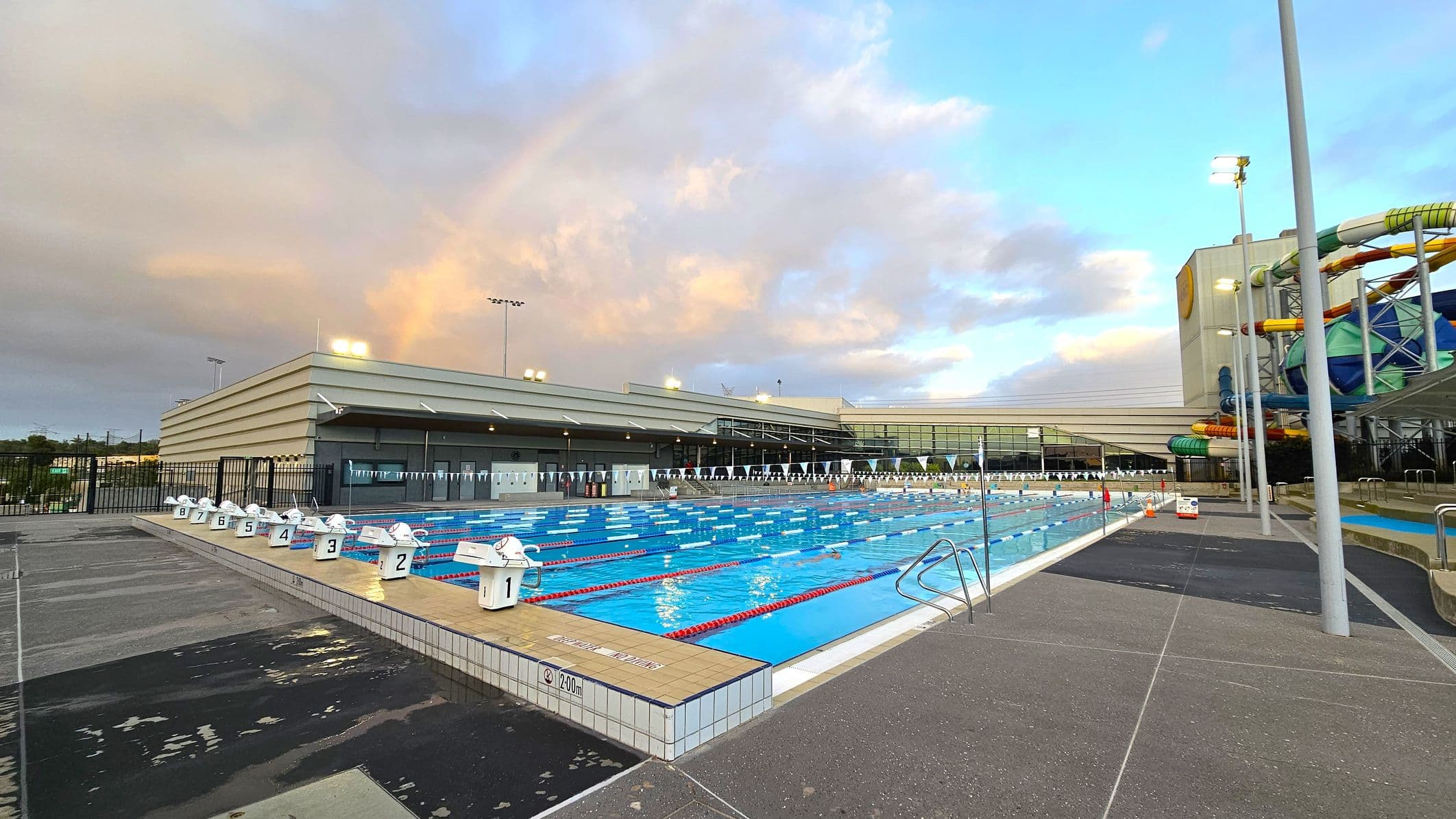 An outdoor swimming pool with eight lanes, each marked with a starting block numbered 1 to 8. The pool is surrounded by a tiled deck. On the right, there is a large water slide structure with colourful tubes. In the background, a modern building with large windows and a flat roof is visible. The area is well-lit with tall floodlights. The ground is a mix of grey and black paving.