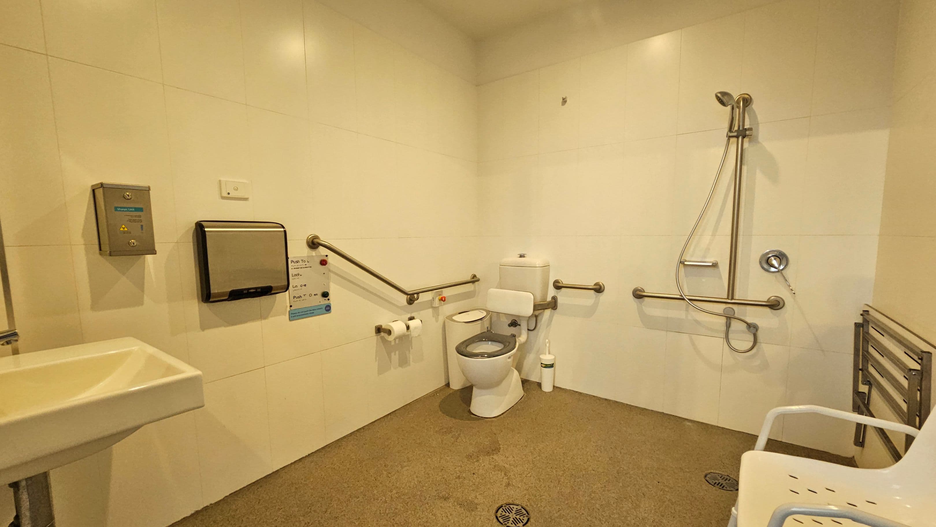 A spacious accessible bathroom with white tiled walls and a grey non-slip floor. On the left, a small white sink is mounted on the wall. In the centre, a toilet with support rails on both sides and a toilet brush nearby. Above the toilet, a paper towel dispenser and a control panel are visible. On the right, a shower area with a handheld showerhead and additional support rails. A white plastic chair with armrests is positioned near the shower.