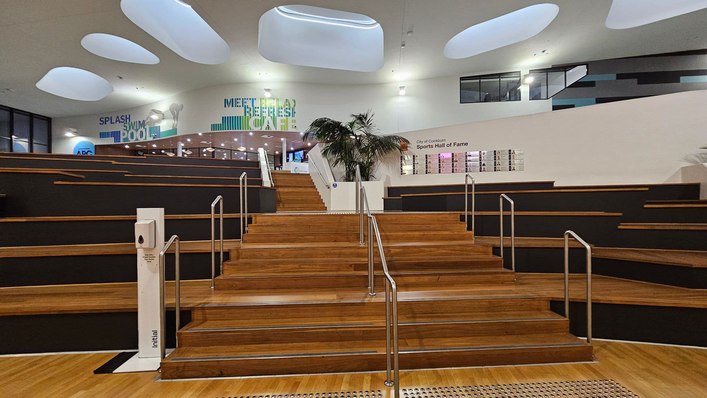 Wooden steps with metal railings lead up to a platform. On the left and right, tiered seating with dark cushions is visible. In the background, a wall displays the "City of Cockburn Sports Hall of Fame" with photos. Above, the ceiling features large, oval-shaped light fixtures. Signs on the wall read "SPLASH SWIM POOL" and "MEET RELAX REFRESH CAFE". A hand sanitiser station is at the base of the steps, and a potted plant is on the platform.
