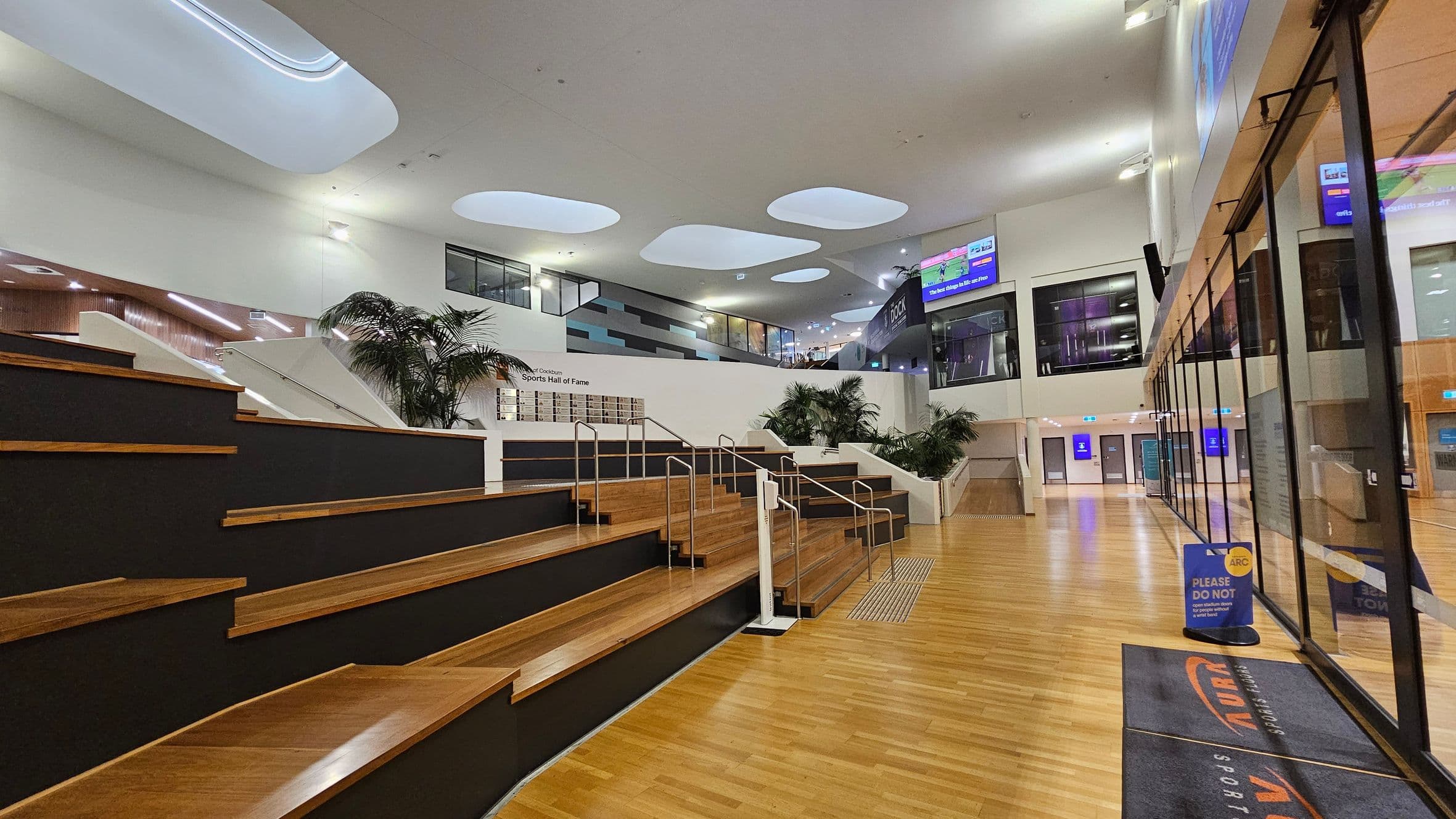 A modern indoor space with wooden tiered seating on the left, featuring metal railings. The floor is wooden, and the ceiling has large, irregularly shaped skylights. On the right, there are glass walls with reflections and a sign on the floor. In the background, there are plants and a wall with a display. Above, digital screens are visible, and doors are at the far end of the space.