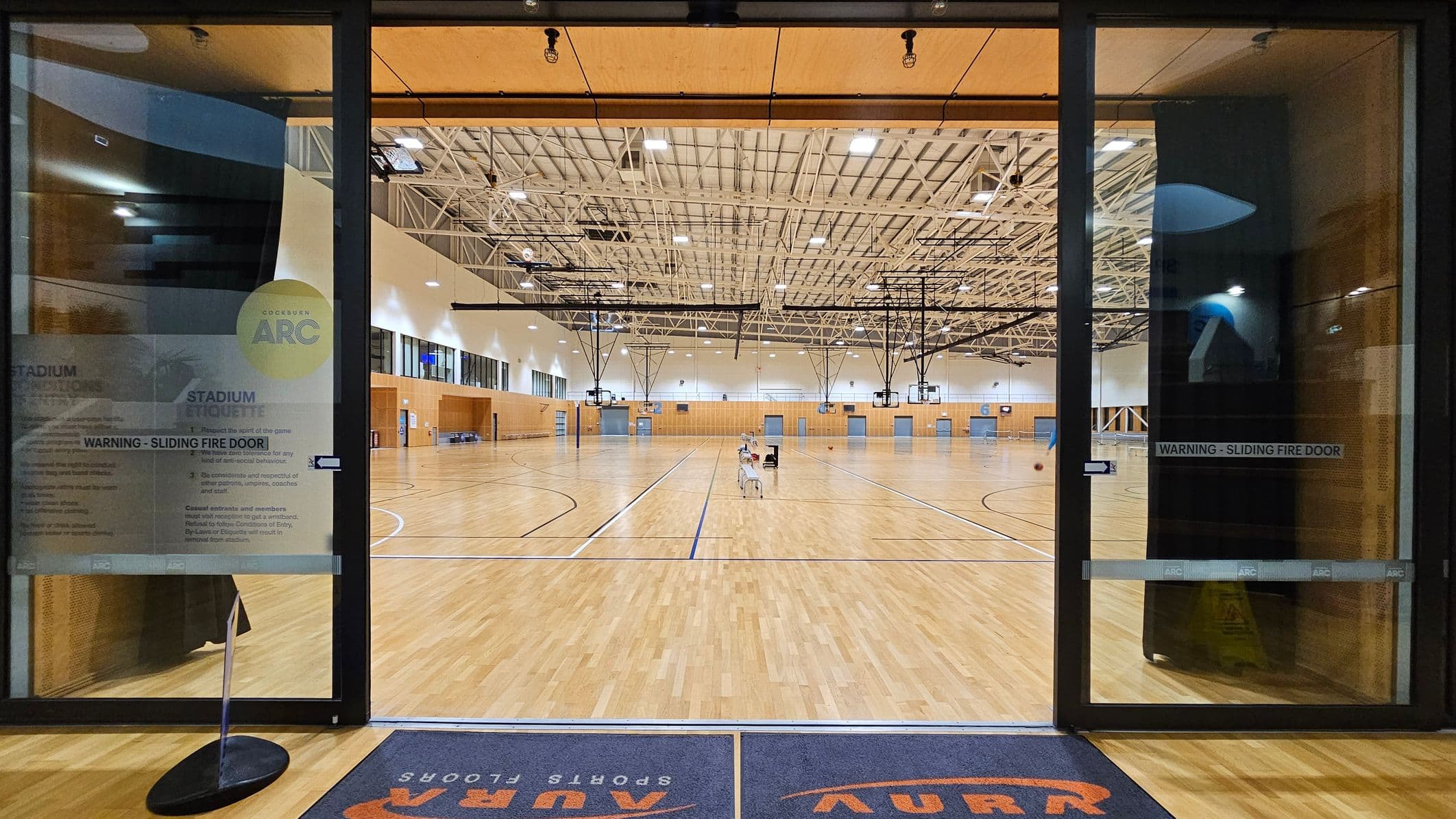A large indoor sports hall with a wooden floor and high ceiling featuring exposed beams and lighting fixtures. In the centre, there are several basketball hoops suspended from the ceiling. The hall is viewed through glass doors with signage, including a warning about a sliding fire door. A blue mat with orange text is on the floor in the foreground. The walls are lined with windows and doors, and there are a few chairs and equipment scattered around the court.