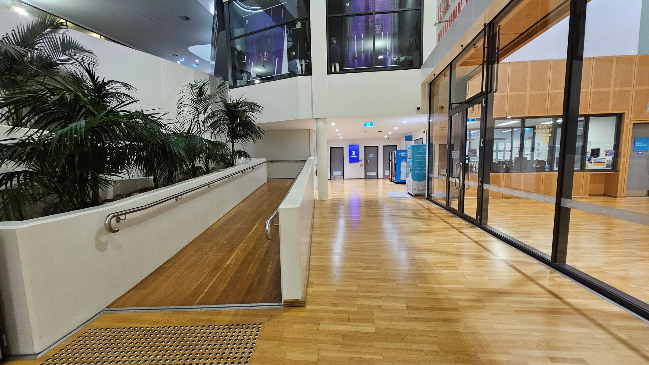 A modern indoor space with a wooden floor. On the left, there's a ramp with a metal handrail, lined with tall green plants. The right side features large glass doors and windows, revealing an interior with wooden panelling and a "Players Register" sign. The ceiling is white with recessed lighting. In the centre, a corridor leads to doors and a digital screen.