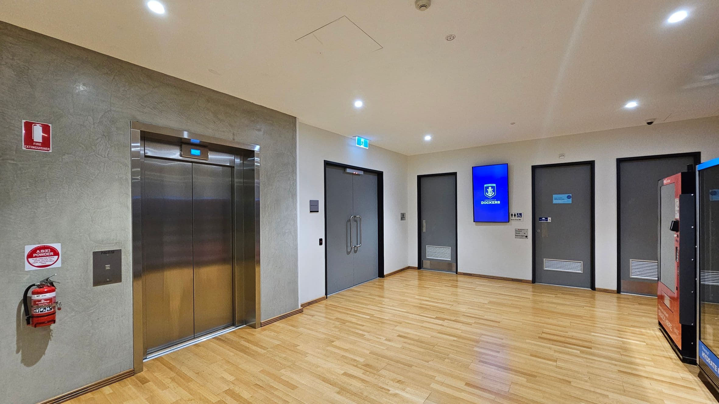 A hallway with a light wooden floor and a white ceiling with recessed lights. On the left, there is a metallic lift with a fire extinguisher mounted on the wall beside it. In the centre, double doors with glass handles are visible. To the right, there are two grey doors and a digital screen displaying a logo. A vending machine is partially visible on the far right.