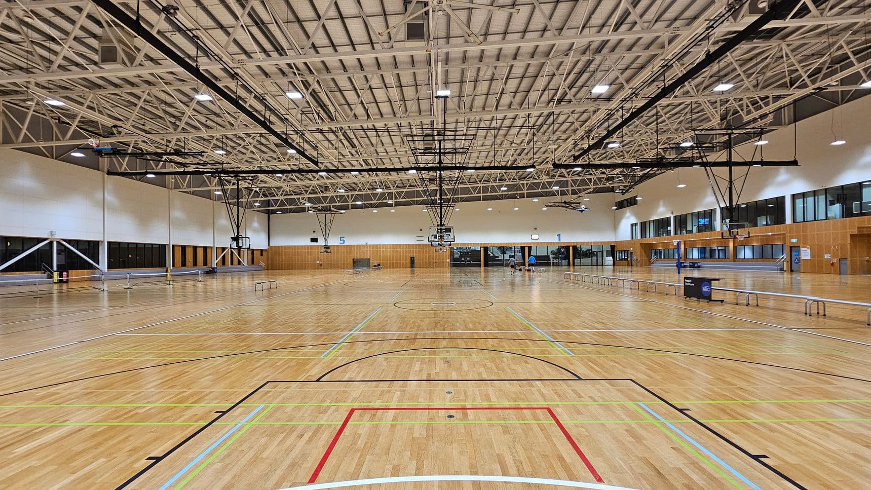 A spacious indoor sports hall with a polished wooden floor marked for various sports. In the centre, basketball hoops are suspended from the high, white truss ceiling. The walls are lined with large windows and wooden panels. On the right, there are seating areas and a few people. The left side features more windows and structural supports. The ceiling is equipped with lights and ventilation fans.