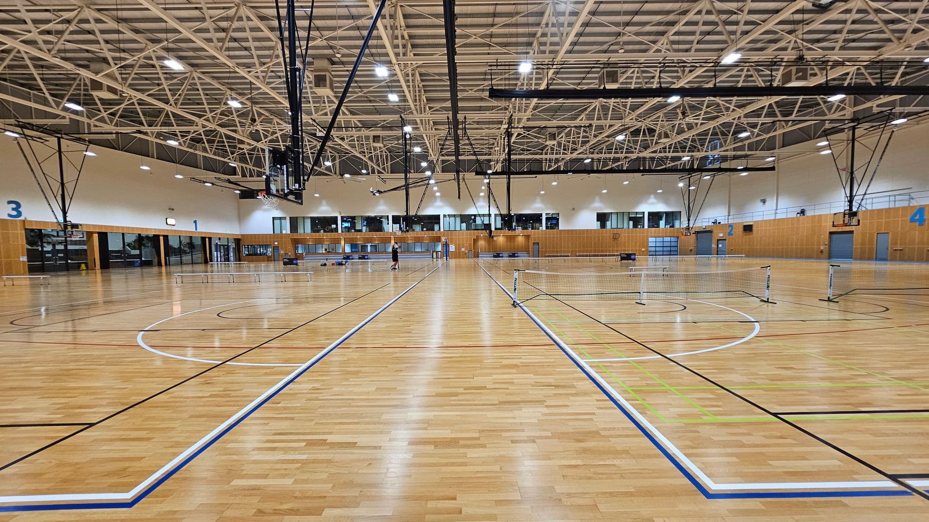 A large indoor sports hall with a polished wooden floor marked for various sports. In the centre, several badminton nets are set up. The ceiling features a complex network of white metal beams with bright overhead lights. On the left and right walls, large numbers "3" and "4" are visible. The background shows a wall with windows and doors, and a few basketball hoops are suspended from the ceiling. A person is visible in the distance near the centre.