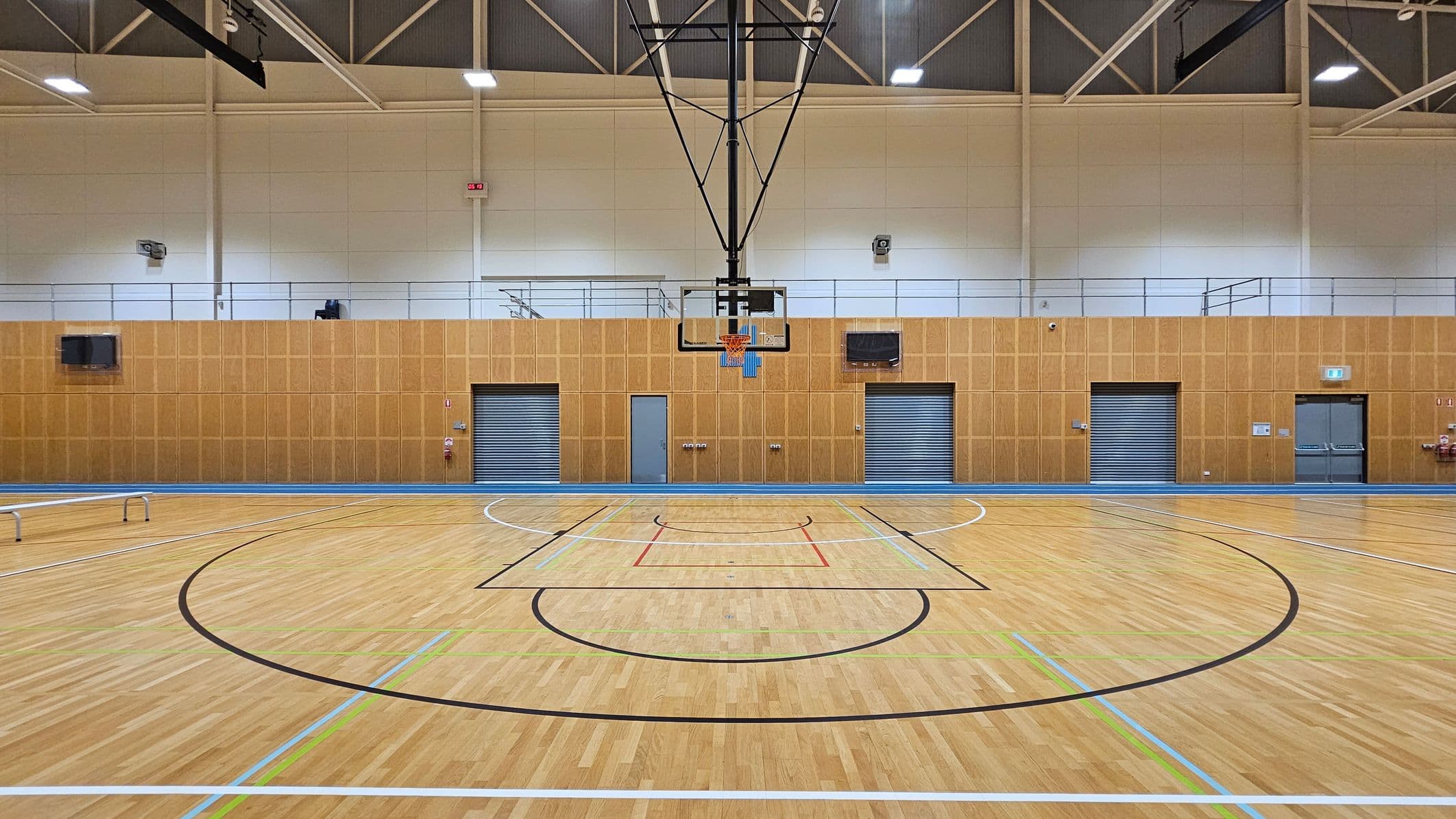 A spacious indoor basketball court with a wooden floor marked with various coloured lines. In the centre, a basketball hoop is suspended from the ceiling. The background features a wooden-panelled wall with three metal roller doors and a single grey door. Above the wall, there is a railing and some equipment. The ceiling is high with exposed beams and bright lights.