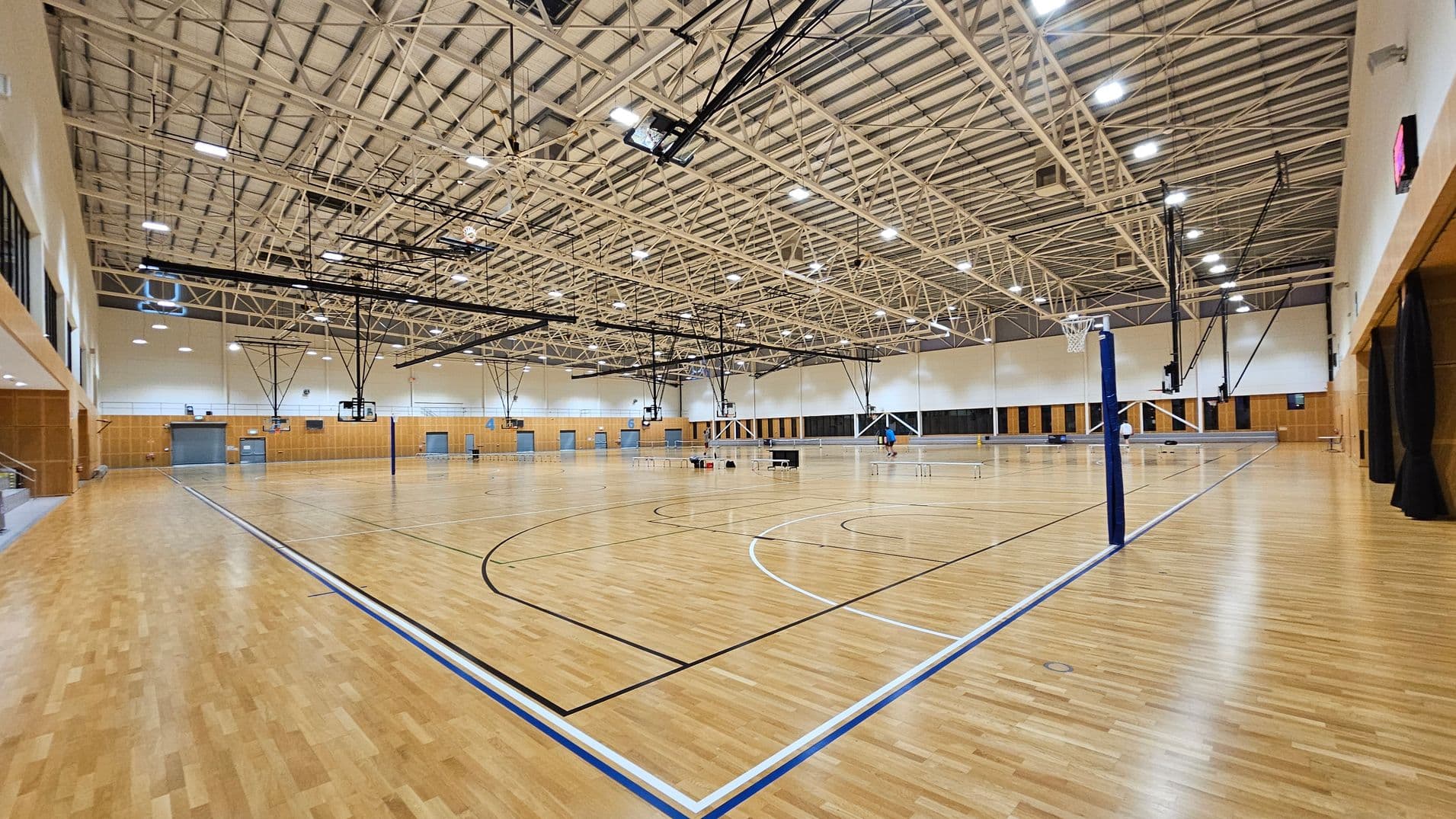 A spacious indoor sports hall with a polished wooden floor marked for basketball. On the left, there are wooden-panelled walls and a small set of bleachers. In the centre, multiple basketball hoops hang from the high, metal-trussed ceiling, which is equipped with bright lights. On the right, a volleyball net is set up. The background features more wooden panels and several doors. The ceiling is high and industrial, with visible beams and lighting fixtures.