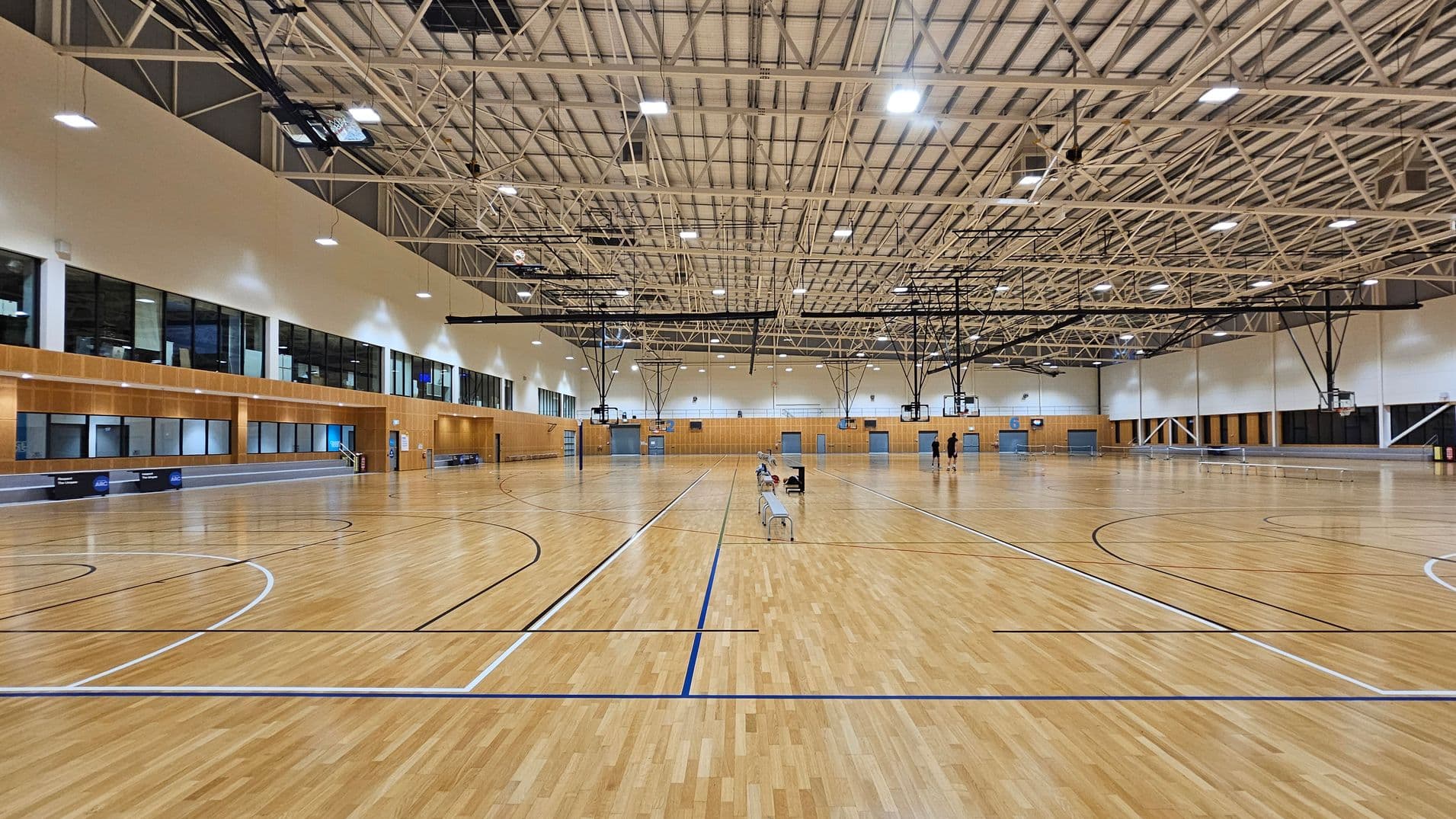 A large indoor sports hall with a polished wooden floor marked for basketball. The ceiling is high with exposed beams and numerous lights. On the left, there are large windows and a wooden-panelled wall. In the centre, a few people are seated on benches, and some are standing. Basketball hoops are suspended from the ceiling. The right side mirrors the left with more windows and a similar wall design. The space is expansive and well-lit.