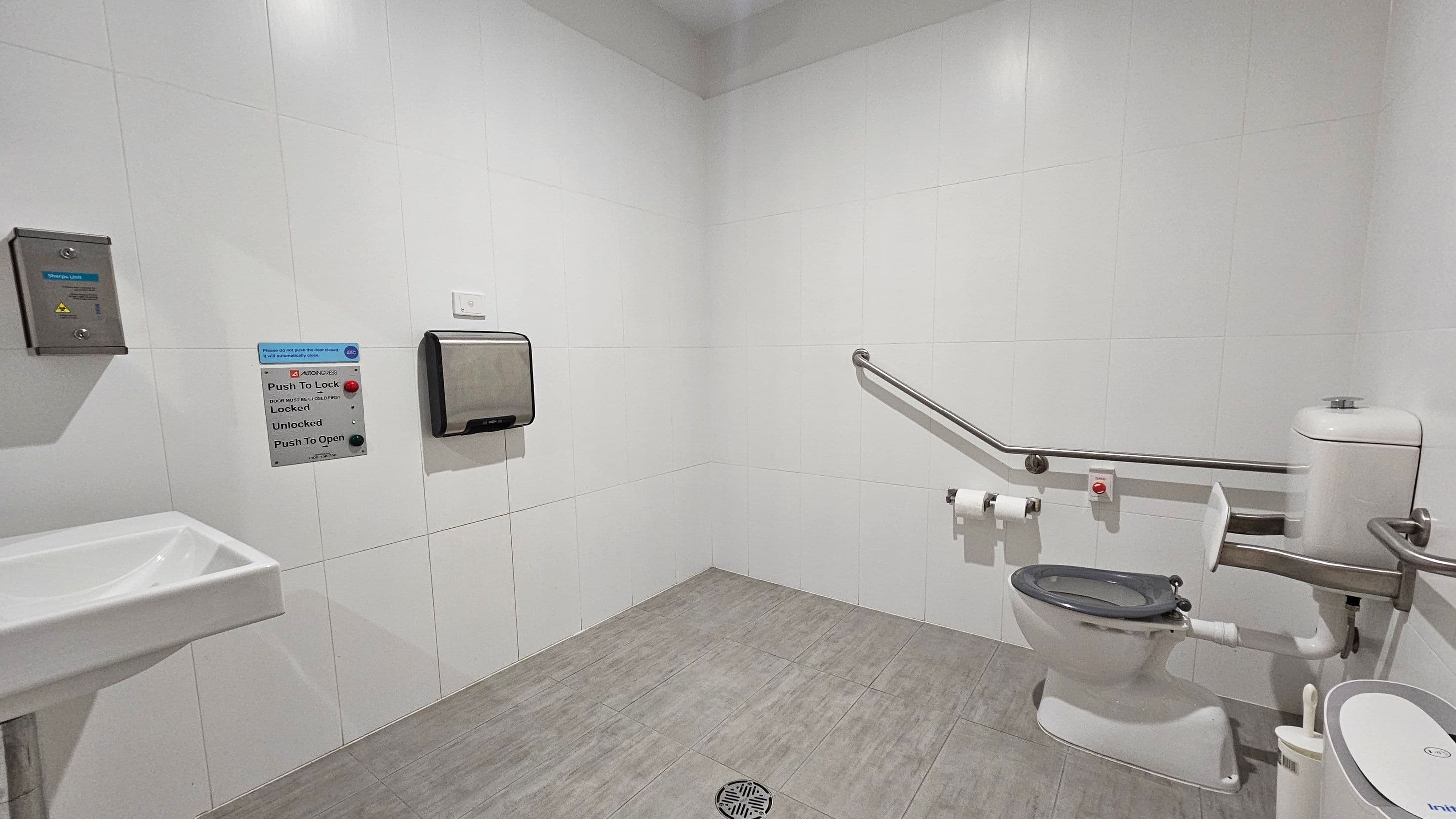 A public accessible toilet with white tiled walls and a grey tiled floor. On the left, a small sink is visible. In the centre, a paper towel dispenser and a sign with buttons for locking and unlocking the door are mounted on the wall. On the right, a toilet with a support rail is present, along with a toilet brush and a bin. A grab bar runs along the wall, and there are two toilet paper holders. A floor drain is near the centre.