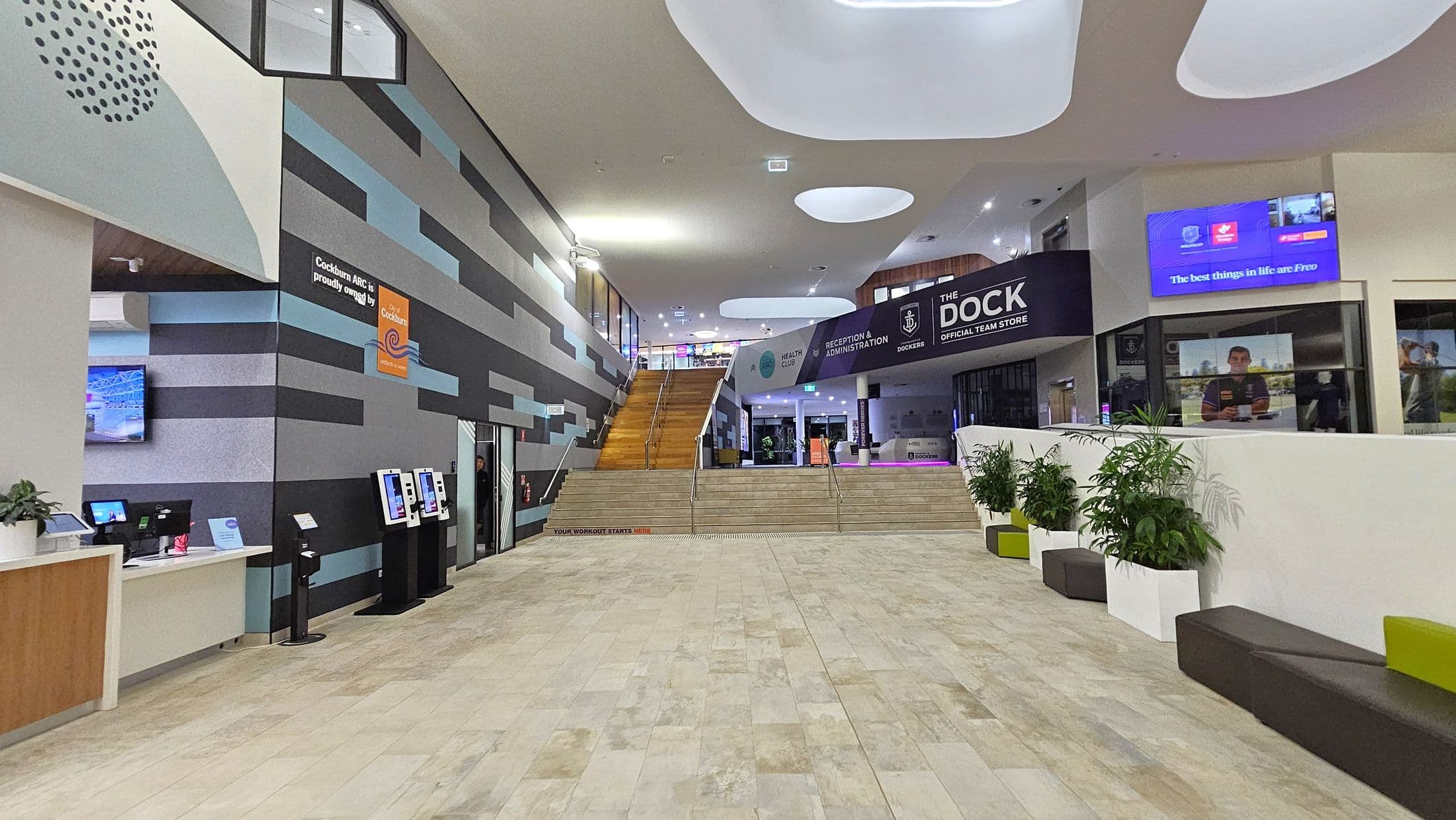 A modern indoor space with a light-coloured tiled floor. On the left, there's a reception desk with digital screens and a wall featuring a geometric pattern in grey, black, and blue. In the centre, a wide staircase leads to an upper level. On the right, there are potted plants in white containers and a seating area with black and green benches. Above, a sign reads "THE DOCK" next to digital displays. The ceiling has large, recessed oval shapes.