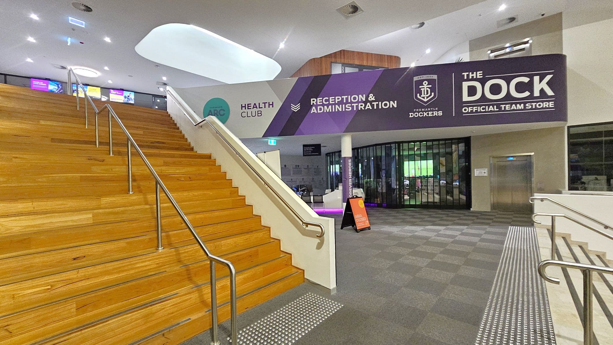 A modern interior with a wide wooden staircase on the left, featuring metal handrails. To the right, a carpeted area leads to a glass-fronted store with a sign reading "THE DOCK OFFICIAL TEAM STORE." Above, a large sign indicates "RECEPTION & ADMINISTRATION" and "HEALTH CLUB." The ceiling is white with recessed lighting, and a curved architectural feature is visible. An elevator is on the far right, and a small sign indicates an area is closed.