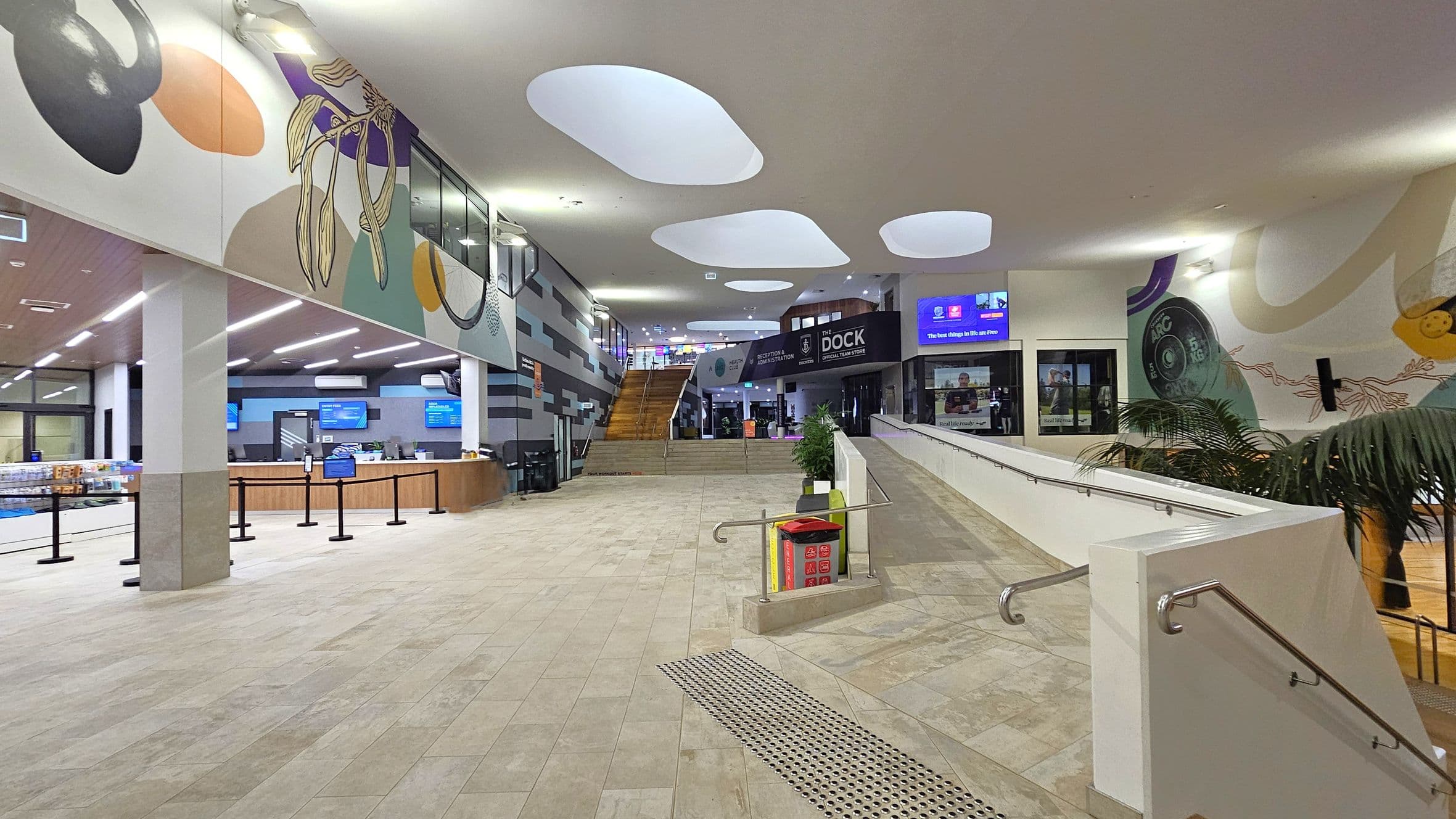 A spacious indoor area with a tiled floor and a ceiling featuring large, irregularly shaped skylights. On the left, there's a reception desk with digital screens above it and a small shop area. In the centre, a wide staircase leads to an upper level. On the right, a ramp with metal railings is lined with potted plants. The walls are decorated with colourful murals, and there are signs for "The Dock" and other facilities.