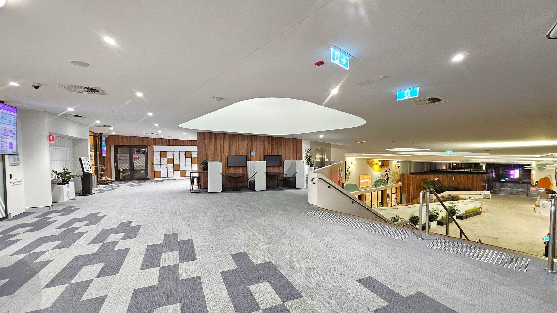 A modern interior space with a grey patterned carpet. On the left, a wall-mounted screen and a fire extinguisher are visible. In the centre, a wooden wall with lockers and two workstations with chairs and screens. The ceiling has recessed lighting and a large, white, curved feature. On the right, a staircase with metal railings leads down to a lower level with more seating and decorative wall art. The area is well-lit with emergency exit signs.