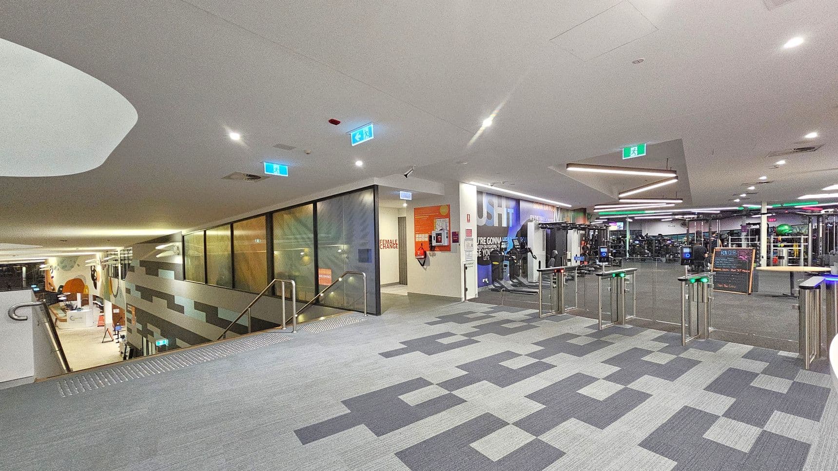 A modern gym interior with a grey patterned carpet. On the left, a staircase with a glass railing leads down to another level. Adjacent to it is a corridor with a sign for a female change room. In the centre, a wall displays motivational text. On the right, turnstiles lead into the main gym area filled with exercise equipment. The ceiling features recessed lighting and geometric light fixtures. Emergency exit signs are visible throughout.