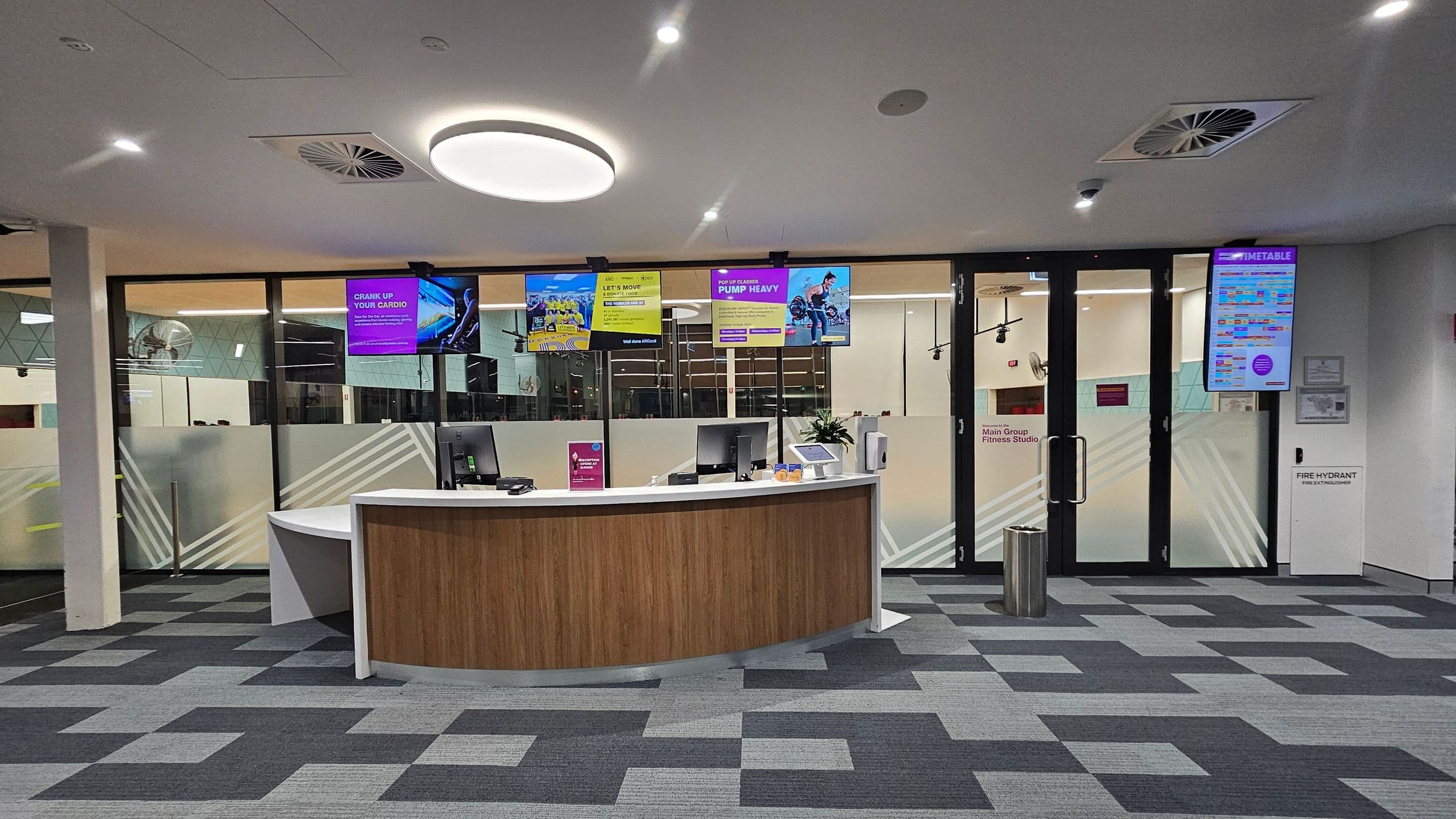 A modern reception area with a curved wooden desk in the centre, equipped with computers and a small plant. Behind the desk, there are glass doors leading to a fitness studio, with several digital screens displaying advertisements and schedules. The floor is covered with a patterned grey carpet, and the ceiling features circular lights and air vents. A sign on the right indicates the location of a fire hydrant.