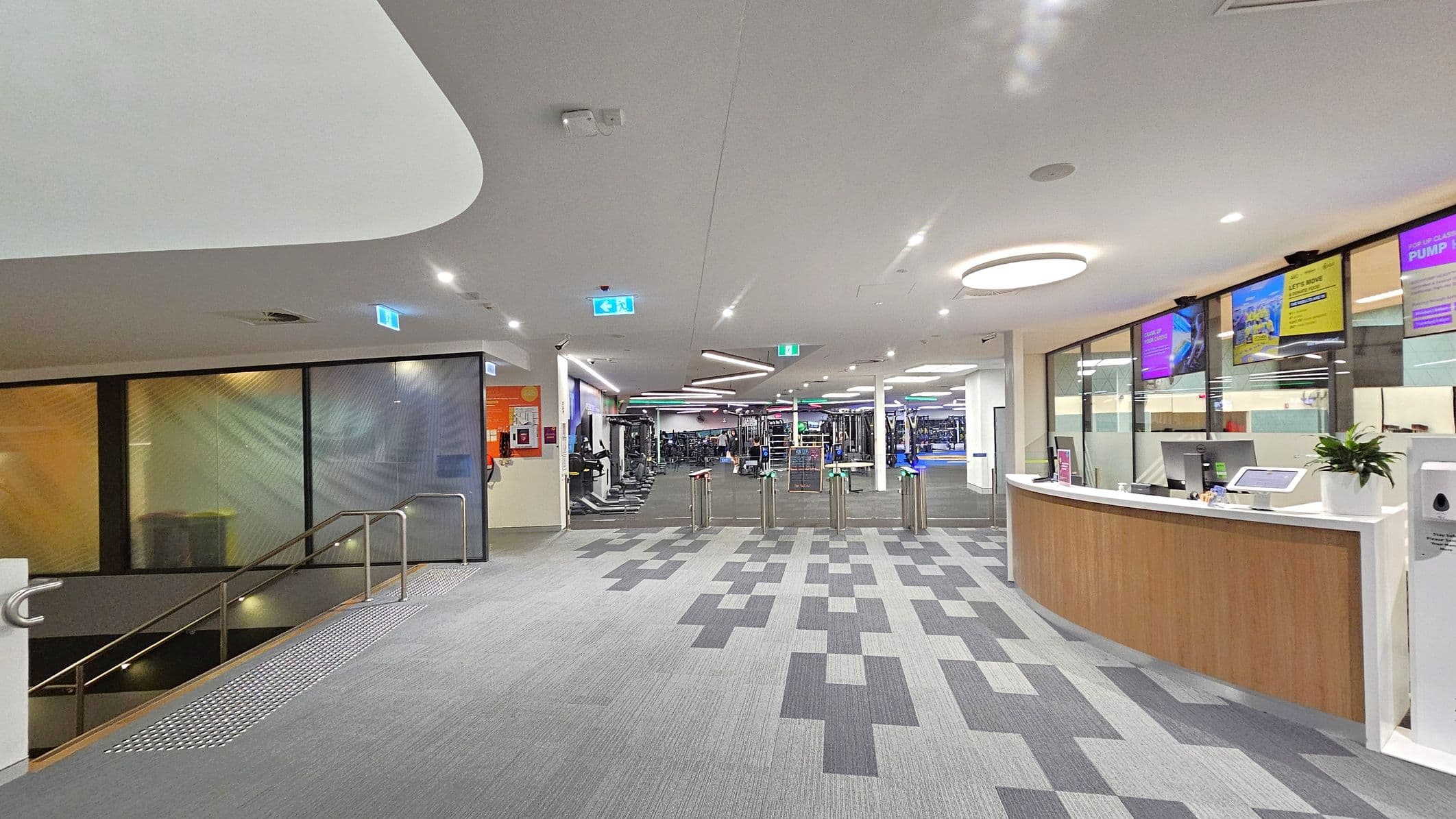 A modern gym entrance with a grey patterned carpet. On the right, there's a curved reception desk with a computer, a plant, and digital screens above displaying information. In the centre, turnstiles lead to the gym area filled with equipment. On the left, a staircase with a metal railing descends, and colourful panels line the wall. The ceiling is white with circular and linear lights.
