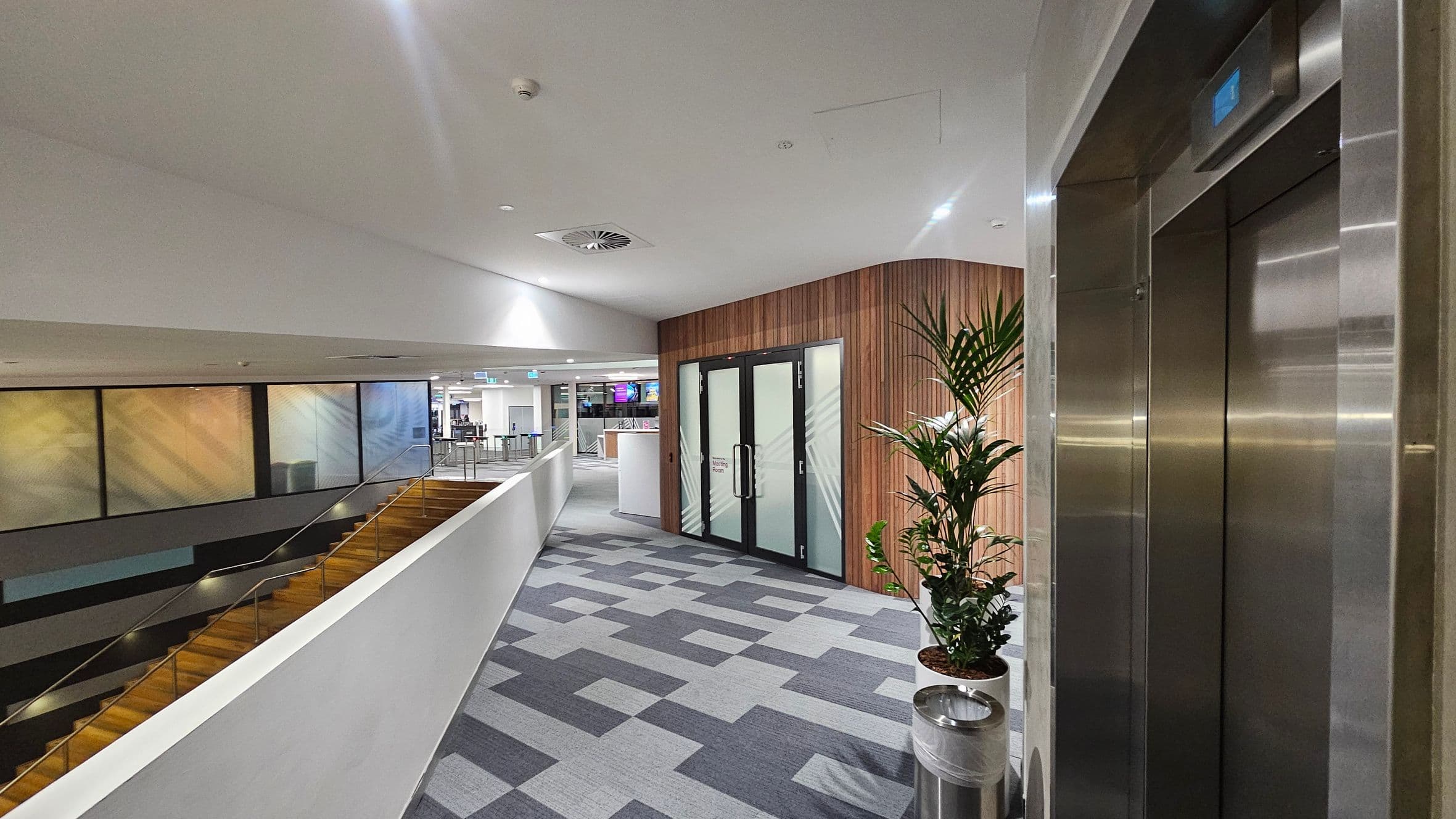 A modern indoor corridor with a grey and purple patterned carpet. On the right, there is a stainless steel lift with a digital display above. Next to it, a potted plant stands beside a metal waste bin. In the centre, a wooden-panelled wall features glass doors with frosted designs. On the left, a staircase with wooden steps and metal railings descends. The background shows a spacious area with colourful panels and a reception desk. The ceiling is white with recessed lighting and air vents.