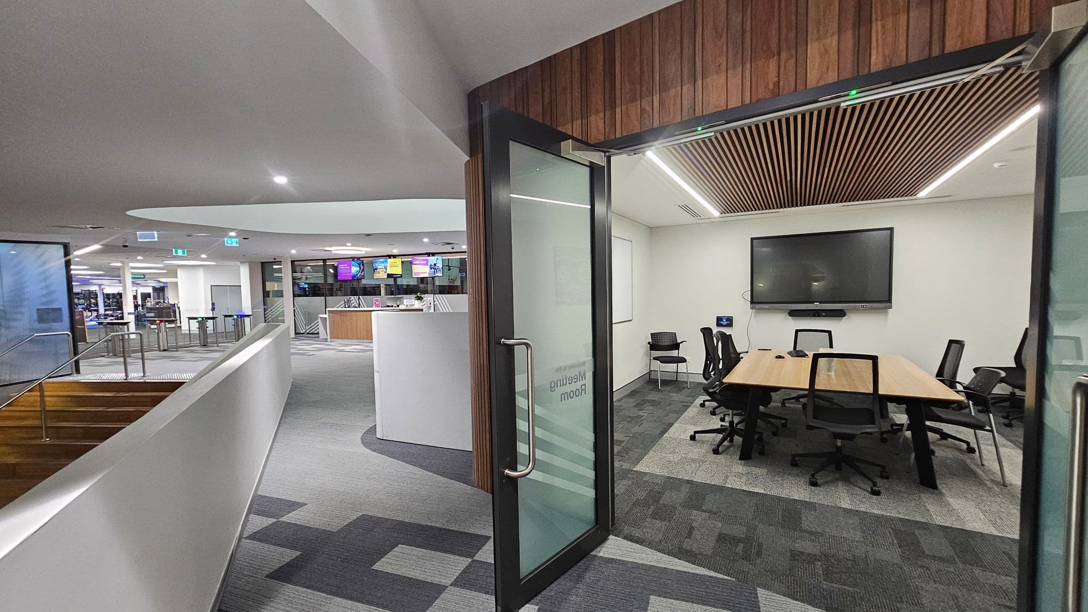 A modern office space with a meeting room on the right. The meeting room has a wooden table surrounded by black chairs, a large screen on the wall, and a slatted wooden ceiling. The door is glass with a metal handle. On the left, there is a corridor with a white railing and stairs leading down. The background shows an open area with a reception desk and digital screens. The floor is carpeted with a geometric pattern, and the ceiling is smooth with recessed lighting.