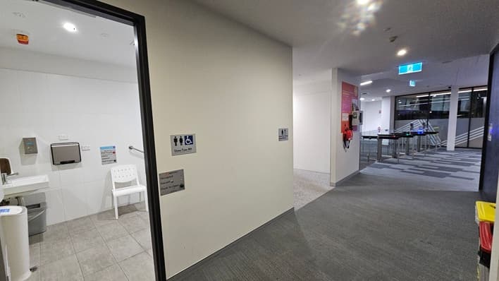 On the left, there's an open door leading to a unisex accessible toilet with a white chair, sink, and paper towel dispenser visible. The floor is tiled. In the centre, a light-coloured wall displays signs indicating the toilet's accessibility. On the right, a hallway with a grey carpet leads to a glass entrance with turnstiles. The ceiling has recessed lighting, and a blue exit sign is visible. A red fire extinguisher is mounted on the wall near the entrance.
