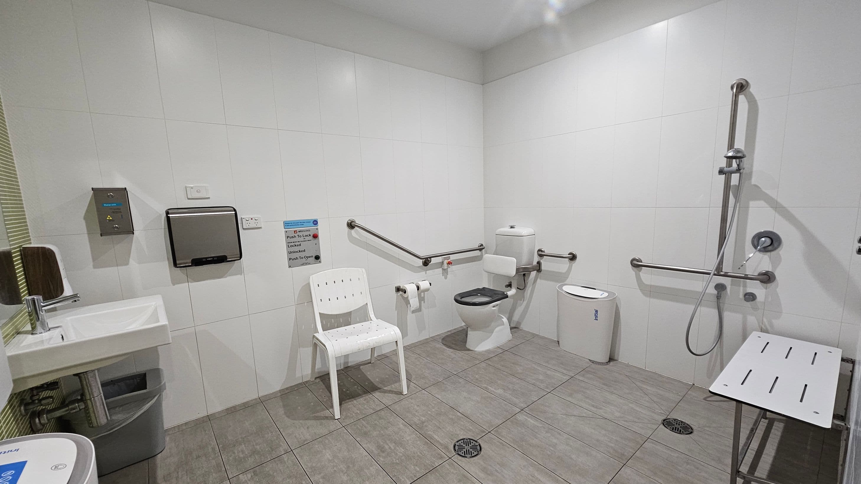A spacious, accessible bathroom with white tiled walls and grey tiled flooring. On the left, a sink with a soap dispenser and a waste bin below. A white chair is positioned near the centre. The right side features a toilet with support rails and a toilet paper holder. A shower area with a handheld showerhead and additional support rails is on the far right, along with a foldable shower seat. A nappy disposal bin is also present near the toilet.