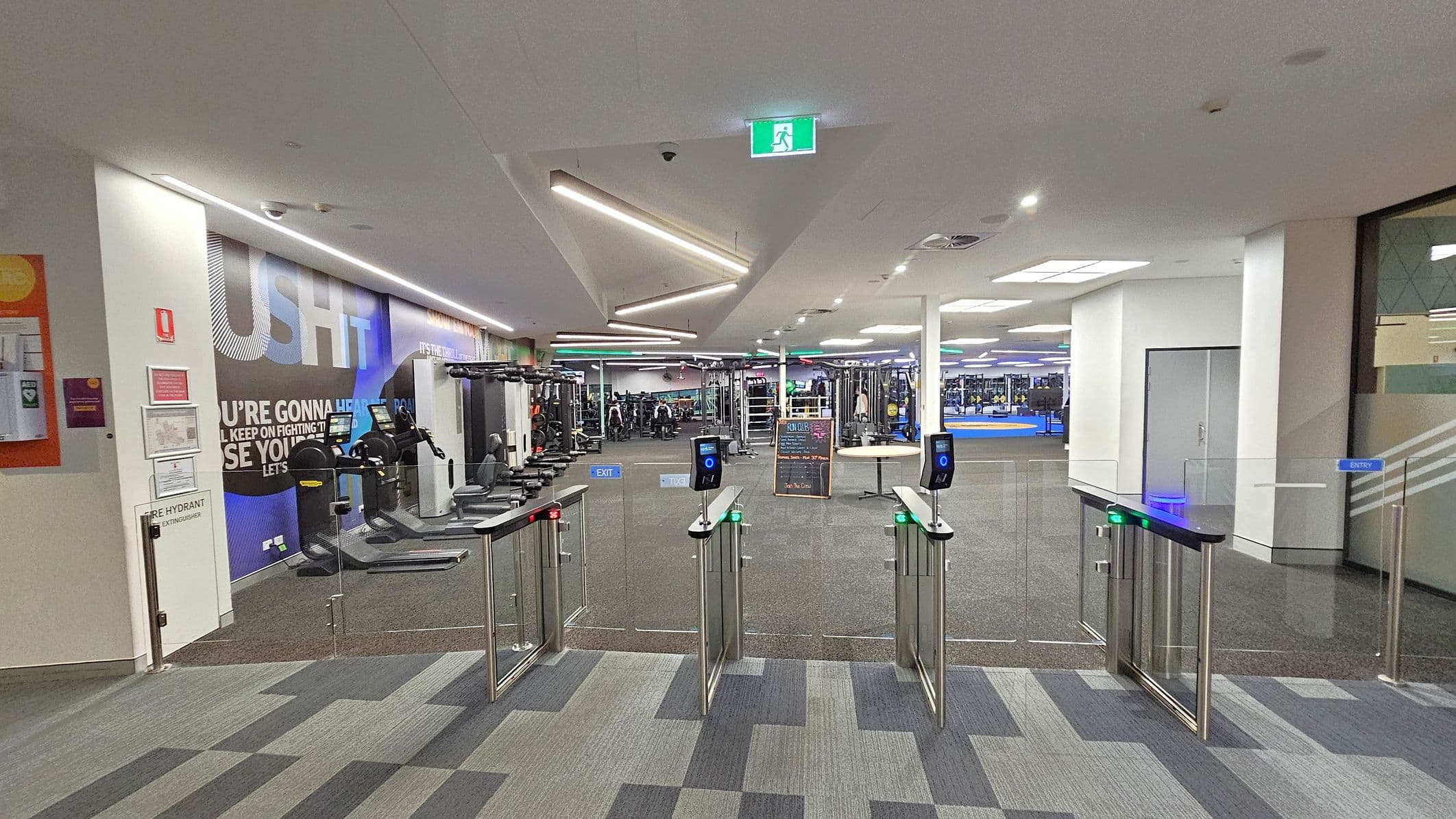 A gym entrance with turnstiles in the foreground, featuring card readers with blue lights. To the left, there's a wall with motivational text and exercise equipment like treadmills. In the background, the gym area is visible with various fitness machines and a chalkboard displaying information. The ceiling has modern, angular lighting fixtures, and the floor is carpeted with a geometric pattern. An emergency exit sign is mounted on the ceiling.