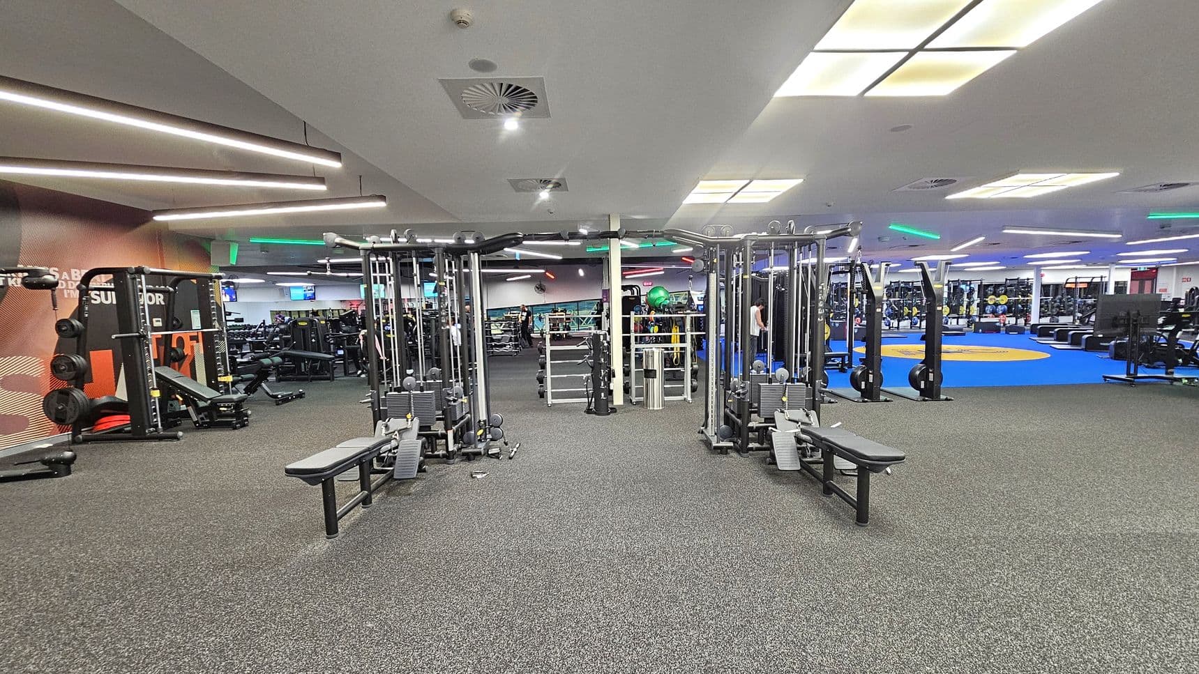 A spacious gym interior with various exercise equipment. In the centre, there are two cable machines with benches. To the left, a squat rack and weightlifting area are visible. On the right, there is a blue mat area with additional equipment like rowing machines. The floor is covered with grey carpet, and the ceiling features bright fluorescent lights and ventilation panels. The gym is well-lit and organised, with a modern design.