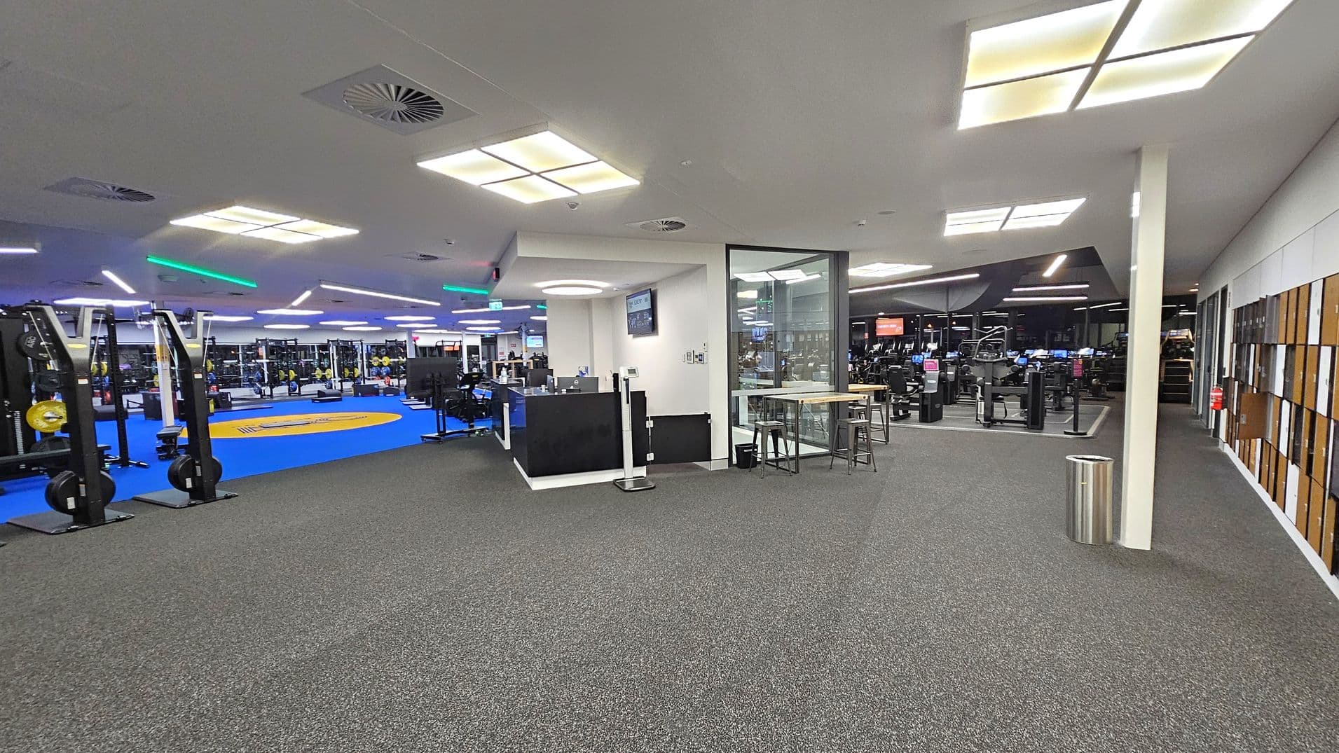 A spacious gym interior with a grey carpeted floor. On the left, there are exercise machines and a blue mat area with weights. In the centre, a reception desk is visible with a screen above it. To the right, there is a glass-walled room with a high table and stools. Further right, more gym equipment is visible, along with a wall featuring wooden panels. The ceiling has square lights and ventilation panels.