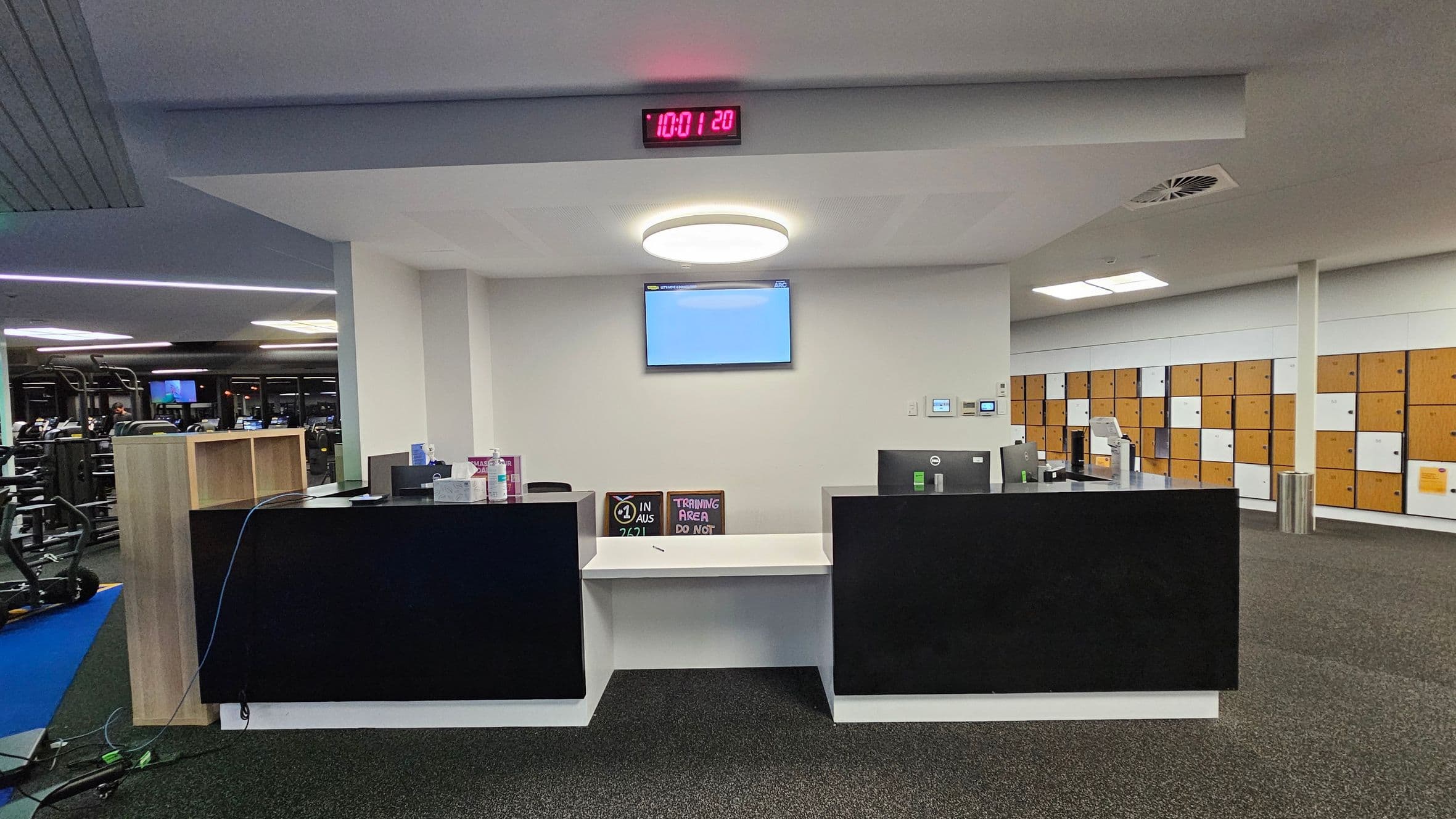A gym reception area with a black counter in the centre, featuring a computer monitor and various items like a sanitiser bottle. Above the counter, a digital clock displays the time. A circular ceiling light is positioned above, with a wall-mounted screen beneath it. To the right, there are rows of wooden lockers with white doors. On the left, gym equipment is visible, including treadmills and exercise machines. The floor is carpeted in dark grey.
