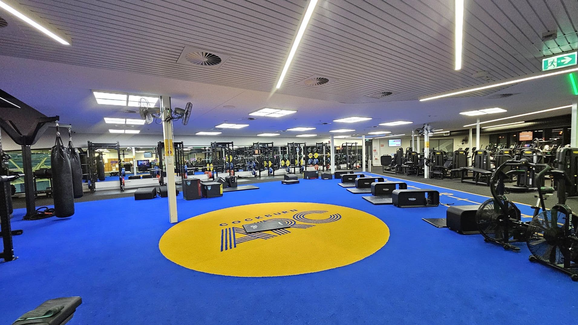 A spacious gym interior with a blue floor featuring a large yellow circle in the centre with "Cockburn ARC" written on it. On the left, there are punching bags and various gym equipment. In the middle, there are weights and exercise stations. On the right, there are cardio machines like exercise bikes. The ceiling has linear lights and ventilation units. The gym is well-lit with a modern design.