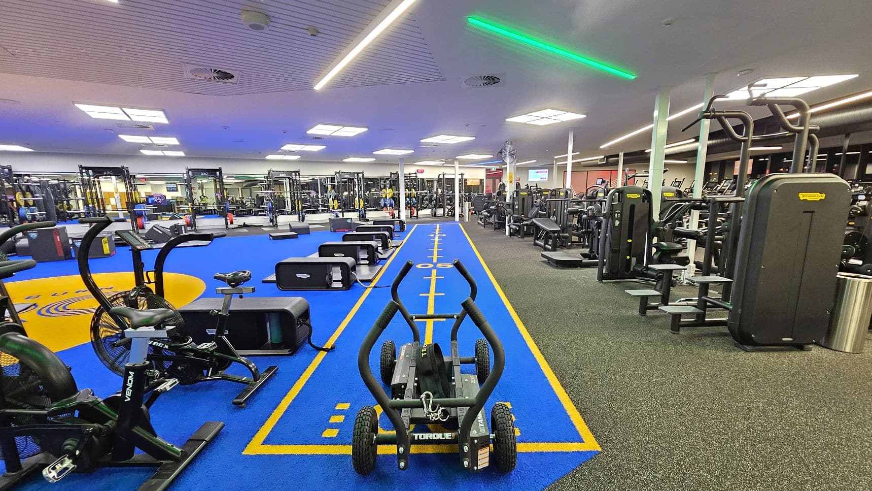 A spacious gym interior with a blue track running down the centre, marked with yellow lines. On the left, there are exercise bikes and weightlifting equipment on a blue floor. On the right, various strength training machines are arranged on a grey floor. The ceiling is lined with white panels and bright lights, with a green strip light adding a modern touch. The gym is well-lit and organised, with equipment neatly placed throughout the space.