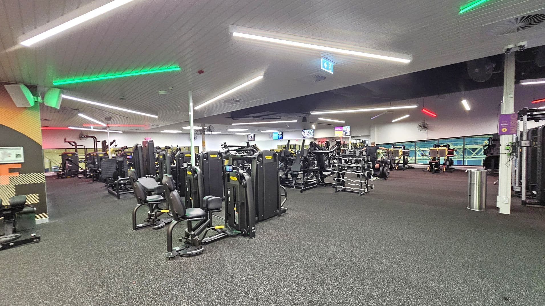 A spacious gym interior with various exercise machines and equipment. On the left, there are weight machines and a wall with a patterned design. In the centre, more machines are arranged in rows, with a rack of dumbbells visible. On the right, additional equipment and a person using a machine can be seen. The ceiling features linear lights with green and red accents, and the floor is covered with dark carpeting. Televisions are mounted on the far wall.