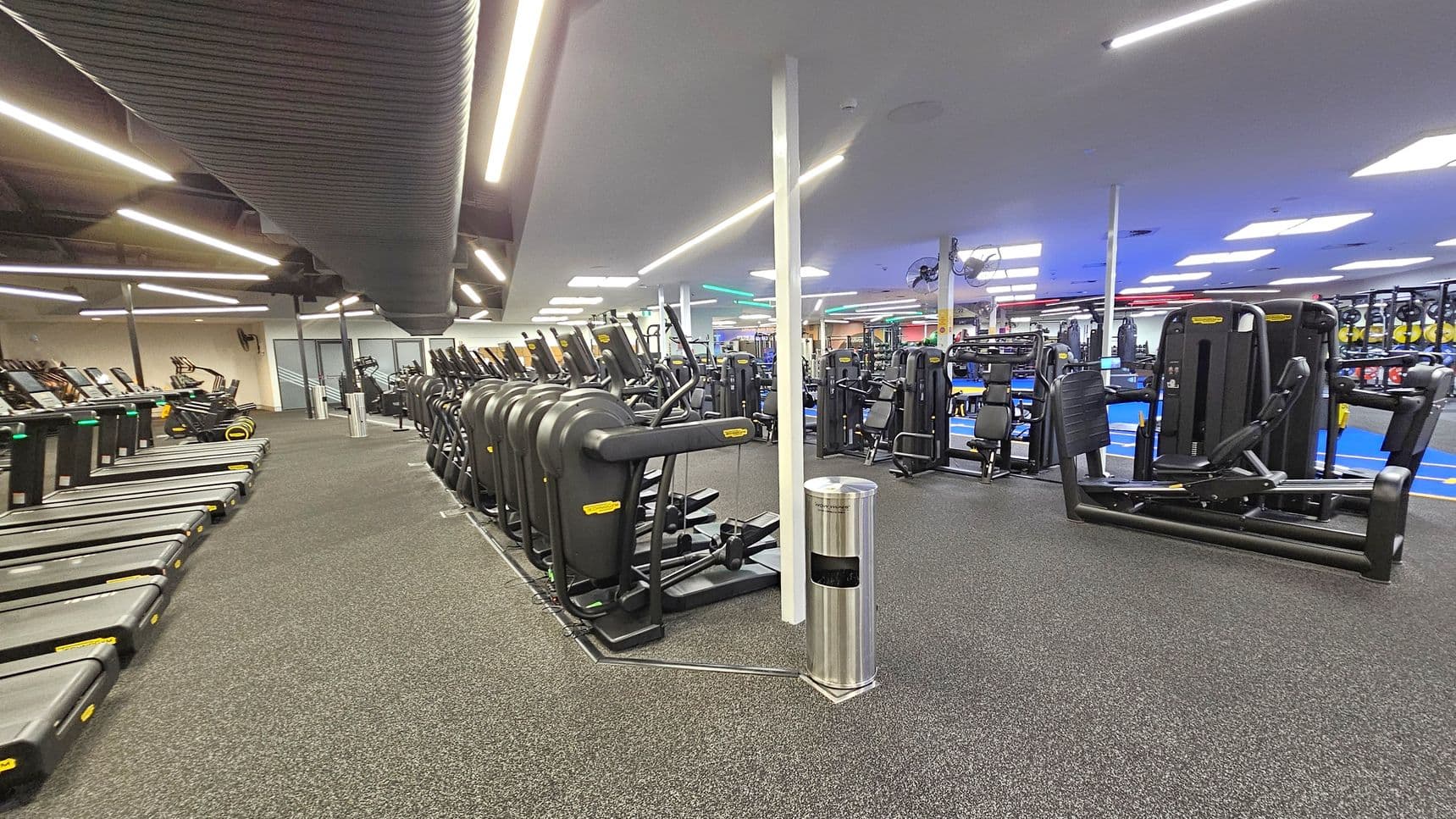 A spacious gym interior with rows of treadmills on the left side, each equipped with digital screens. In the centre, there are several elliptical machines lined up neatly. On the right, various strength training machines are visible. The floor is covered with dark, speckled rubber matting. The ceiling features long, bright fluorescent lights and a large, black ventilation duct running across. A silver rubbish bin is positioned near a white pillar in the middle.