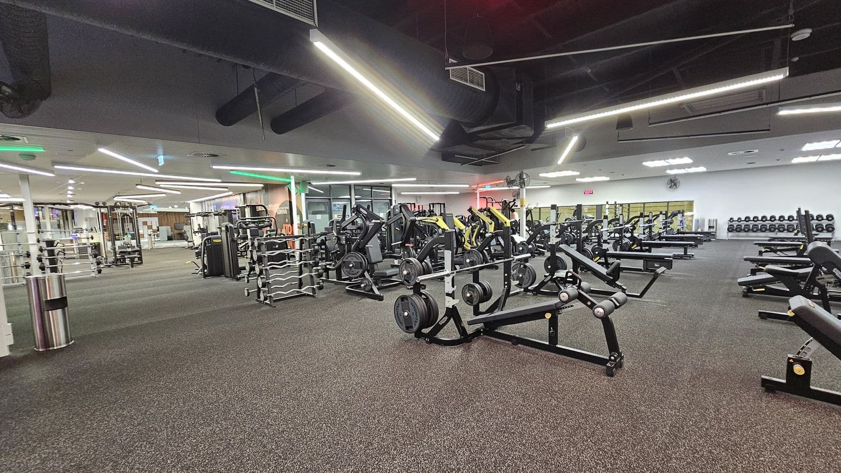 A spacious gym interior with various exercise machines and equipment. On the left, there are racks of weights and a metal bin. In the centre, multiple weightlifting machines and benches are arranged neatly. The right side features more benches and a wall lined with dumbbells. The floor is carpeted in a speckled pattern, and the ceiling has exposed ducts with bright linear lights. The background includes mirrors and glass doors.