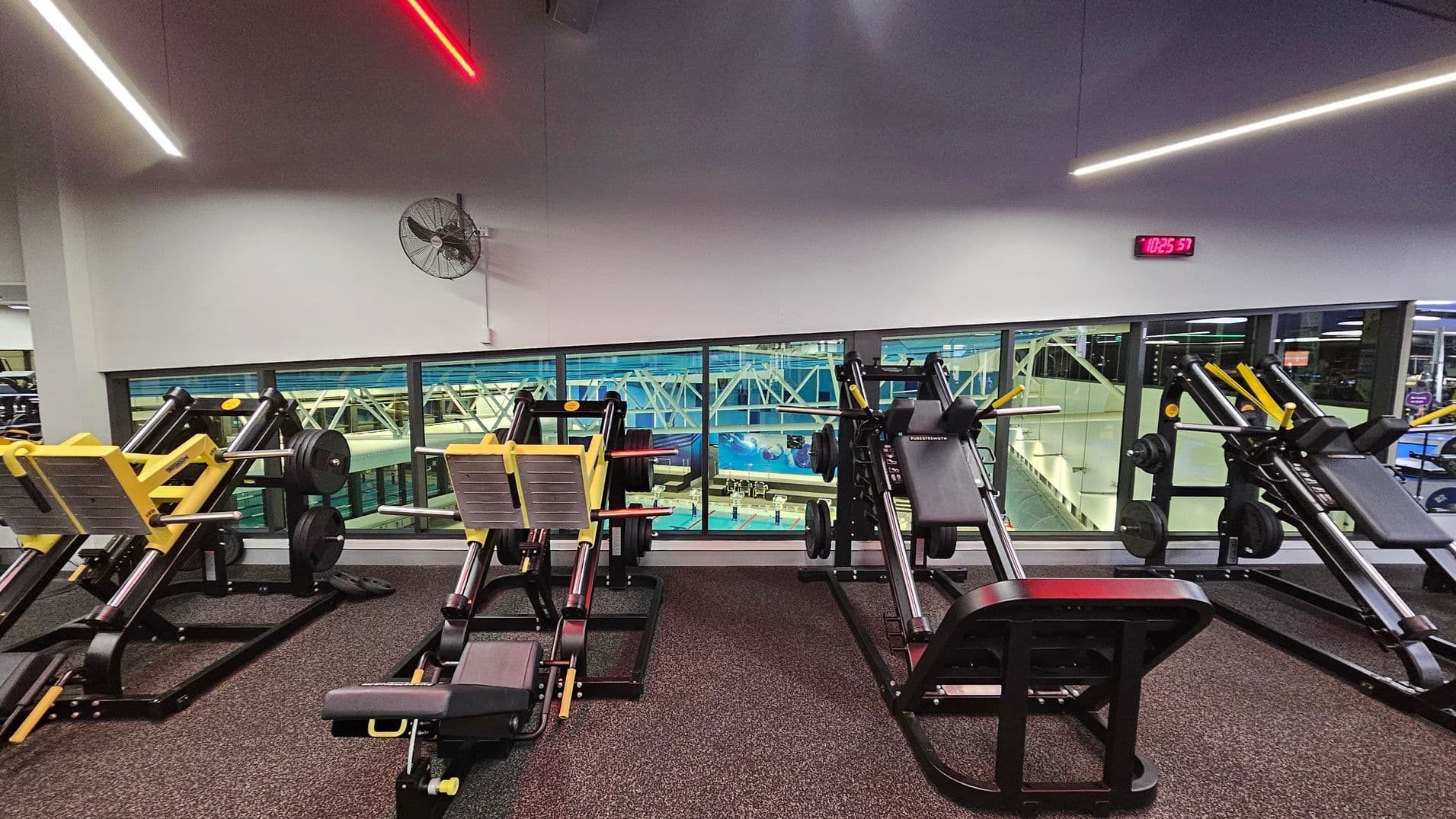 A gym area with several leg press machines featuring yellow and black accents on a dark carpeted floor. The wall behind has a large window overlooking an indoor swimming pool with lanes and blue water. A wall-mounted fan is on the left, and a digital clock displaying "10:25:57" is on the right. The ceiling has linear LED lights in white and red.
