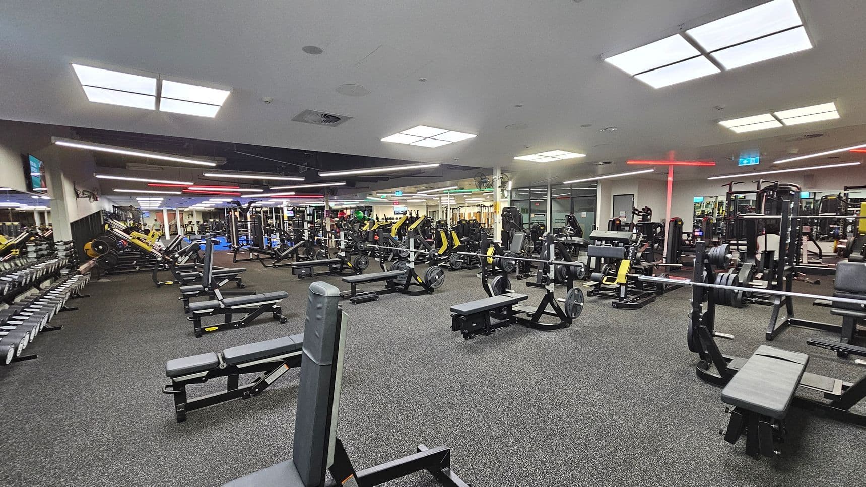 A spacious gym with various exercise equipment. On the left, there are rows of dumbbells and benches. In the centre, multiple weightlifting machines and benches are arranged neatly. The right side features more weight machines and benches. The floor is covered with dark grey carpet, and the ceiling has bright rectangular lights. The background shows mirrors and additional gym equipment.
