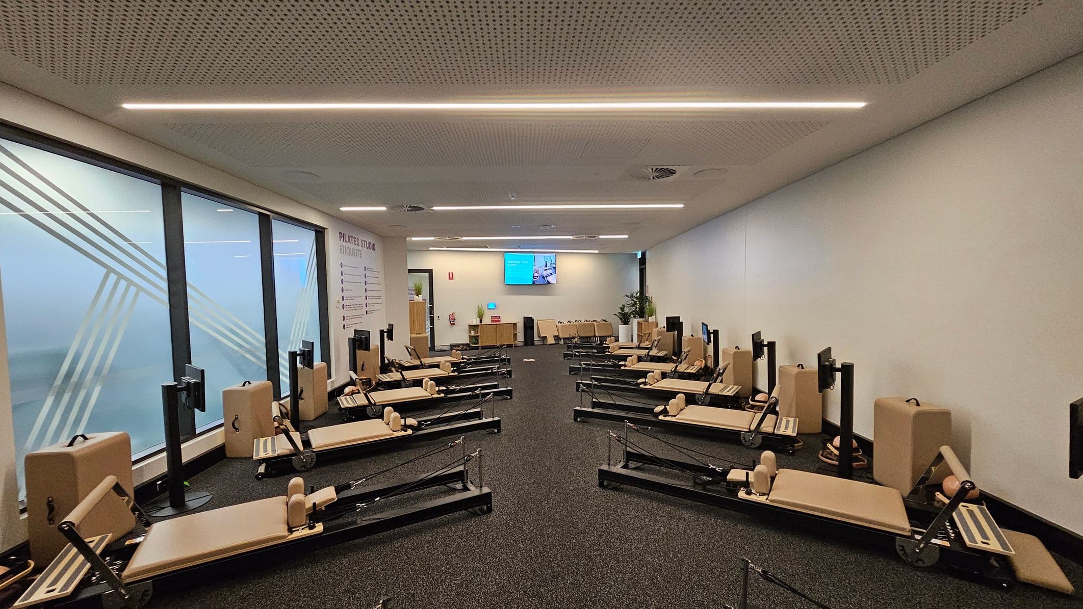 A Pilates studio with multiple reformer machines arranged in two rows on a dark carpeted floor. The left wall features large frosted glass windows with diagonal patterns, while the right wall is plain white. At the back, a wall-mounted screen is visible above a wooden cabinet, with potted plants nearby. The ceiling is perforated with linear lights. A sign on the left wall outlines studio etiquette.