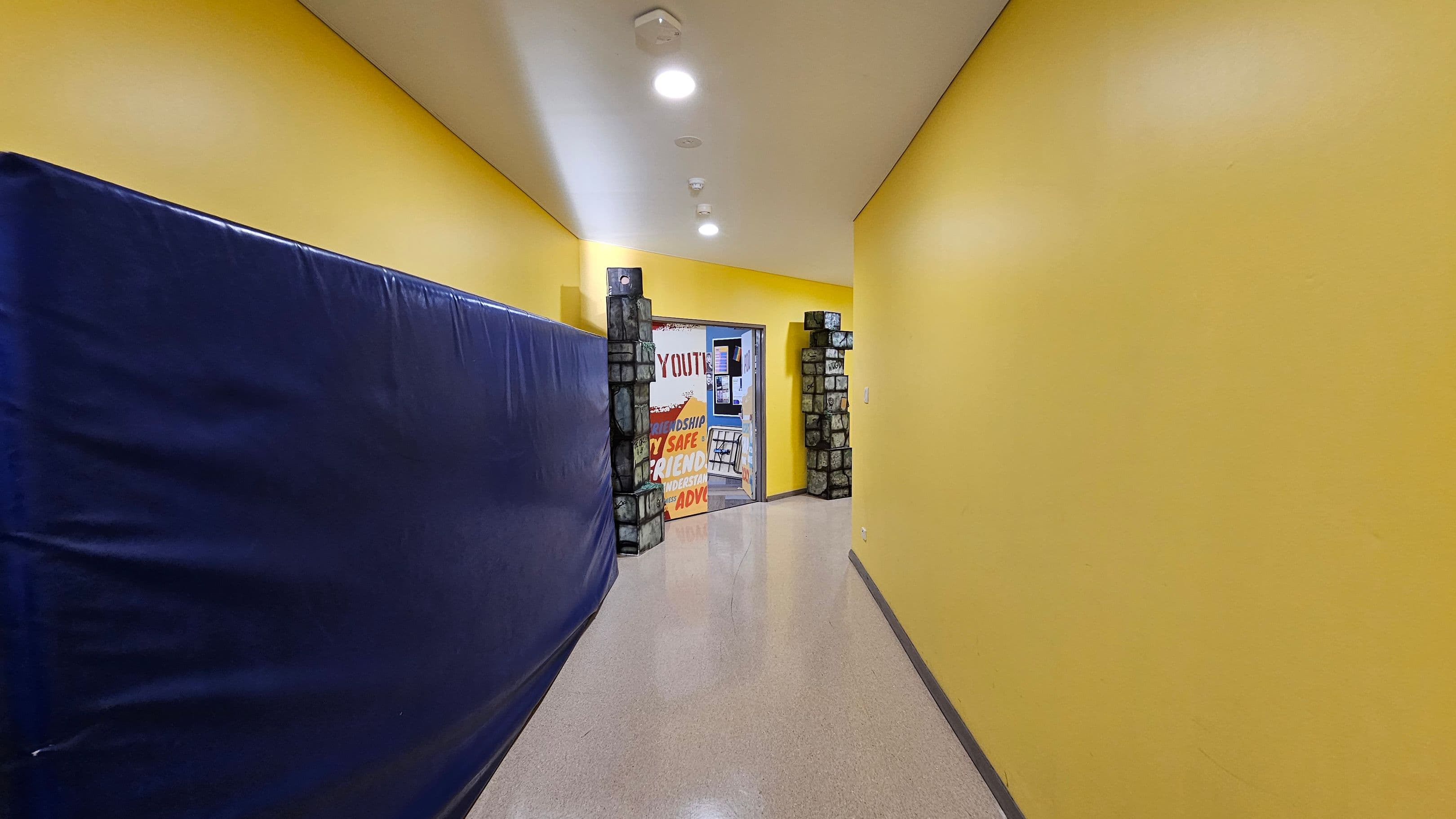 A hallway with bright yellow walls and a glossy floor. On the left, a large blue padded mat covers the wall. At the end of the hallway, there's an open doorway framed by decorative stone-like pillars. Through the doorway, a colourful sign with words like "YOUTH," "SAFE," and "FRIENDSHIP" is visible. The ceiling is white with recessed lighting.