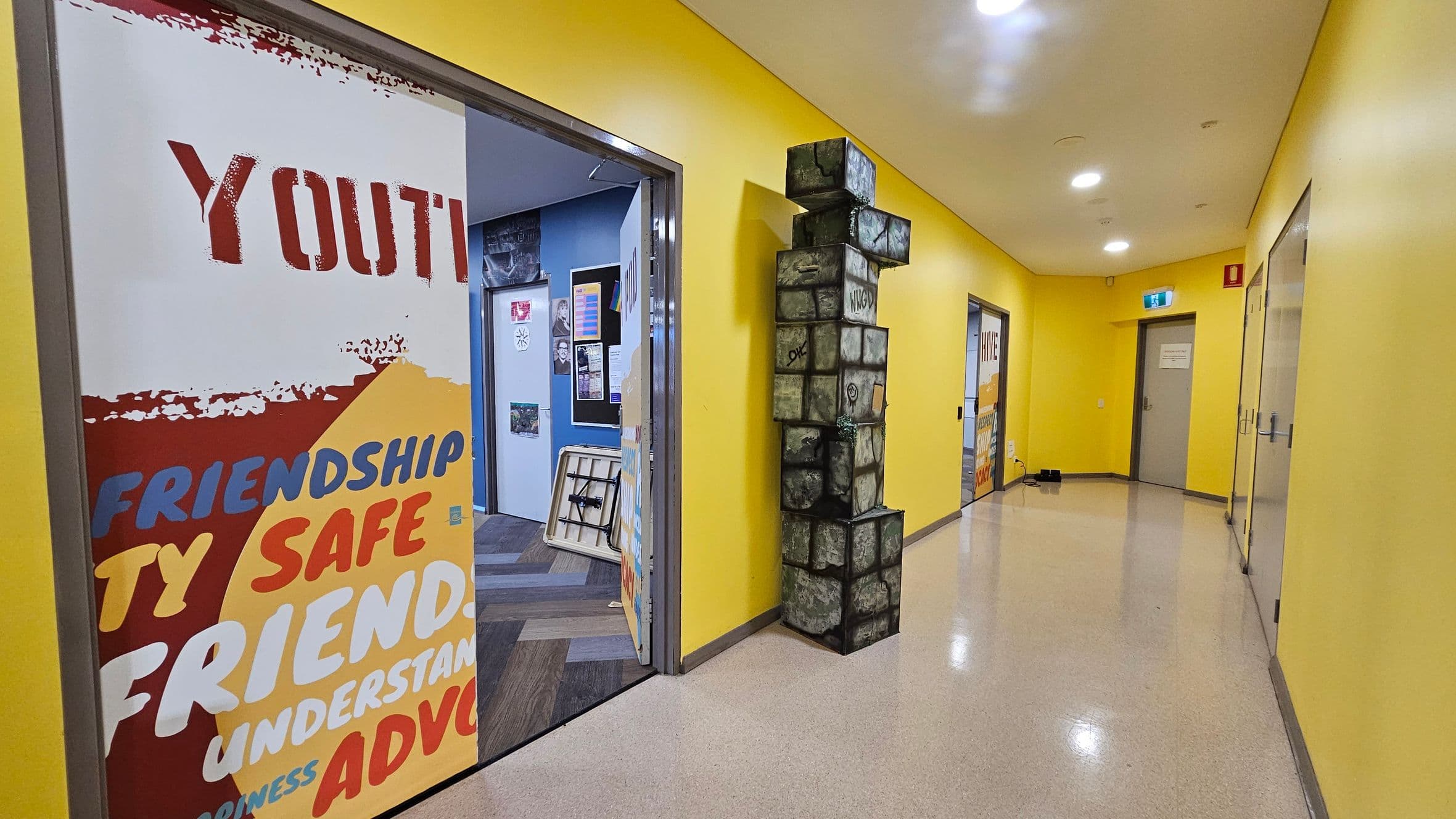 A hallway with bright yellow walls and a shiny, light-coloured floor. On the left, a door is open, revealing a room with blue walls and various posters. A large poster on the door displays words like "YOUTH," "FRIENDSHIP," and "SAFE." In the middle, a stack of cube-shaped blocks with a stone-like appearance is positioned against the wall. On the right, another door is visible, and further down the hallway, an exit sign is mounted above a closed door. Ceiling lights illuminate the space.