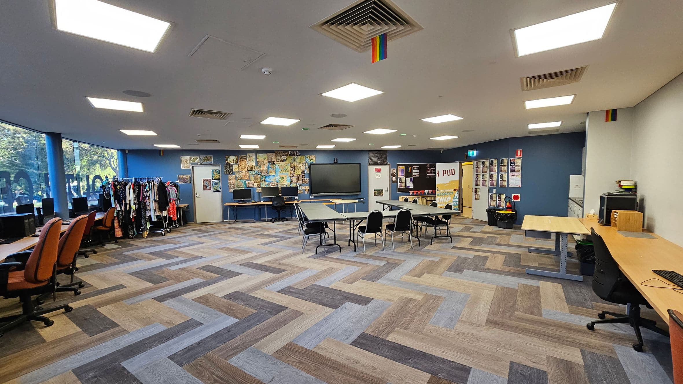 A spacious room with a herringbone-patterned floor in various shades of brown and grey. On the left, there are several orange office chairs in front of computer desks, with a clothing rack nearby. The centre features a large screen on the wall, surrounded by a collage of posters. In the middle, tables and chairs are arranged. On the right, more desks and chairs are visible. The ceiling has bright square lights and a rainbow flag. The walls are blue and white, with more posters and a door.