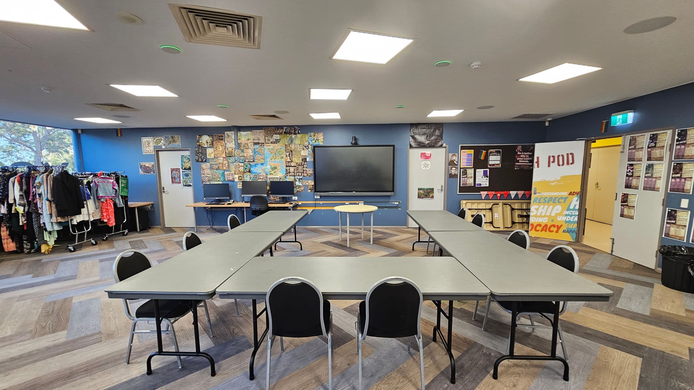 A room with a U-shaped arrangement of tables and chairs in the centre. The floor has a multicoloured wood pattern. The back wall is blue, featuring a large display screen flanked by two computers on a desk. Posters and maps cover the wall. On the left, clothing racks are filled with various garments. On the right, a door with a "POD" sign and a bulletin board with notices. The ceiling has square lights and air vents.