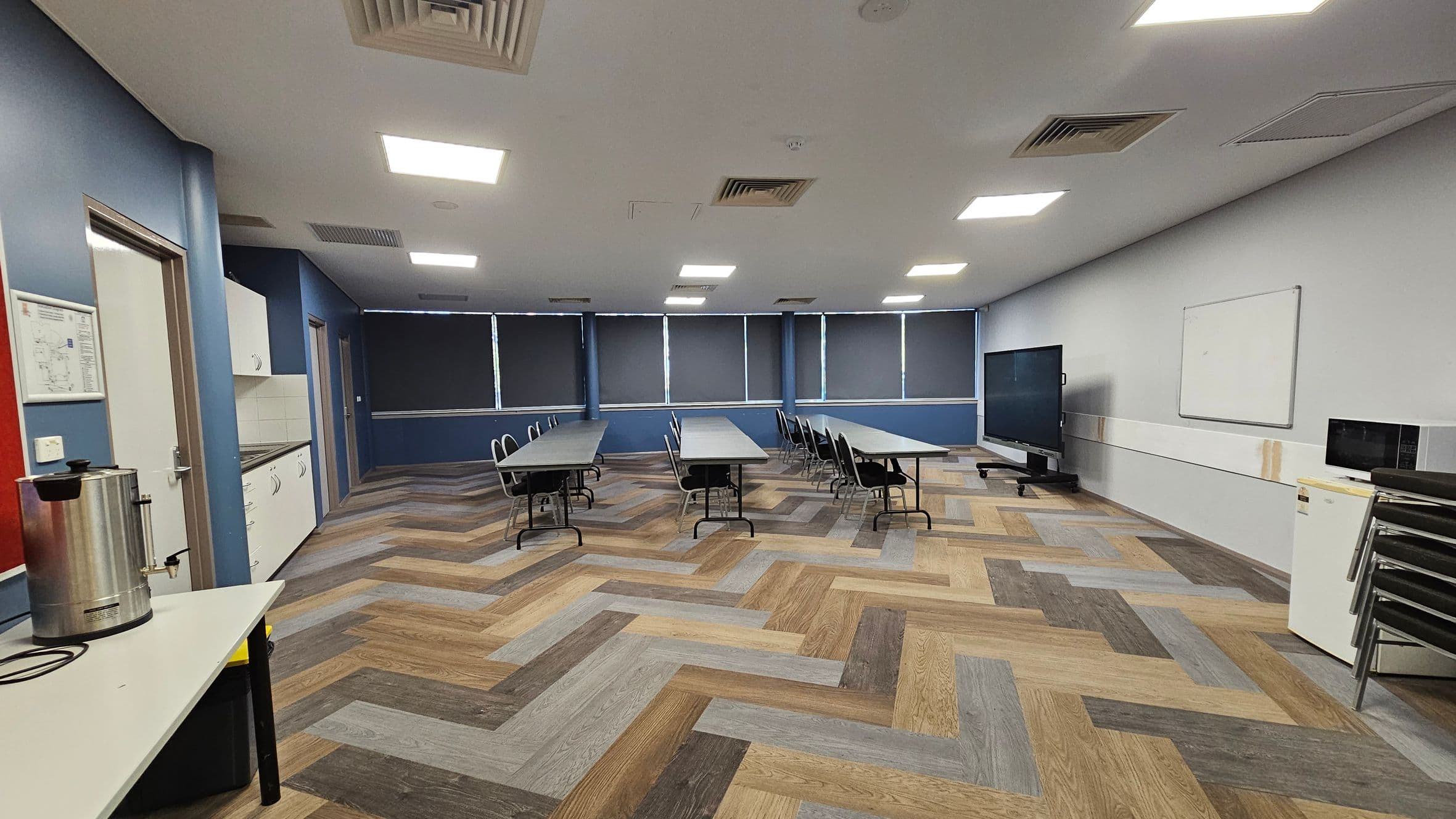 A room with a herringbone-patterned wooden floor and a white ceiling with recessed lights. On the left, there's a counter with a large metal urn and a door. Cabinets and a sink are along the left wall. In the centre, several tables with chairs are arranged in rows. The right side has a large screen on a stand, a whiteboard, and a small fridge with a microwave on top. The windows at the back are covered with dark blinds.