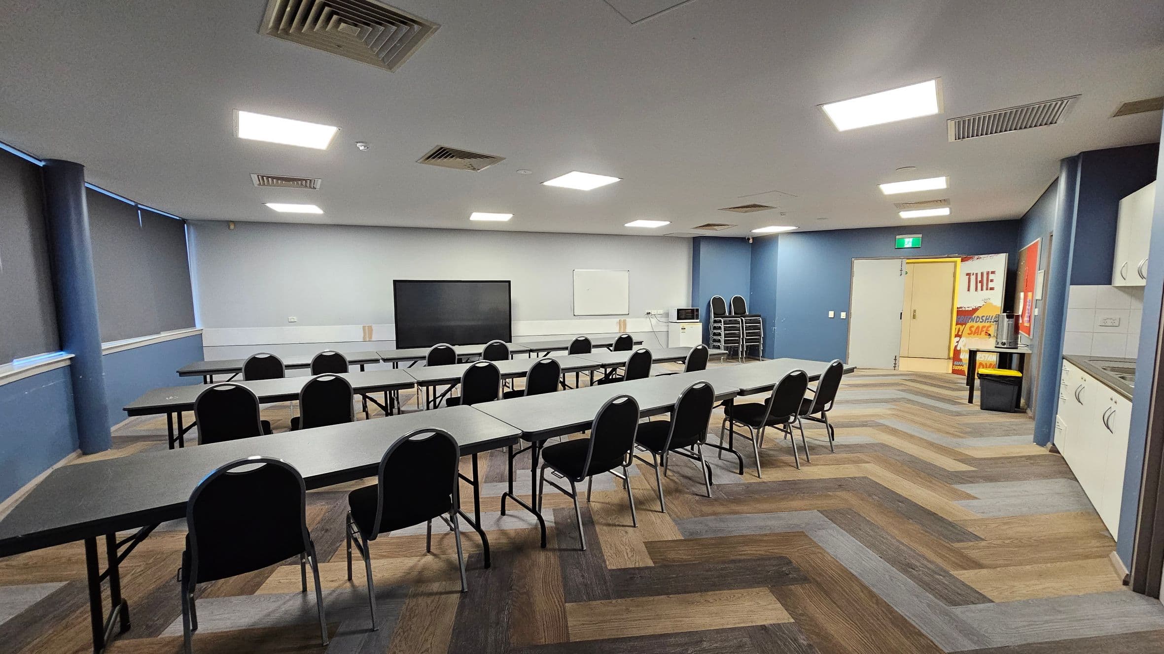 A room with a zigzag patterned wooden floor and a ceiling with recessed lighting and vents. On the left, there are long tables with black chairs facing a large screen and a whiteboard on the wall. The right side has a kitchenette with white cabinets and a stack of chairs. In the background, there's a door with a green exit sign and a poster next to it. The walls are painted blue and grey, and the windows have dark blinds.